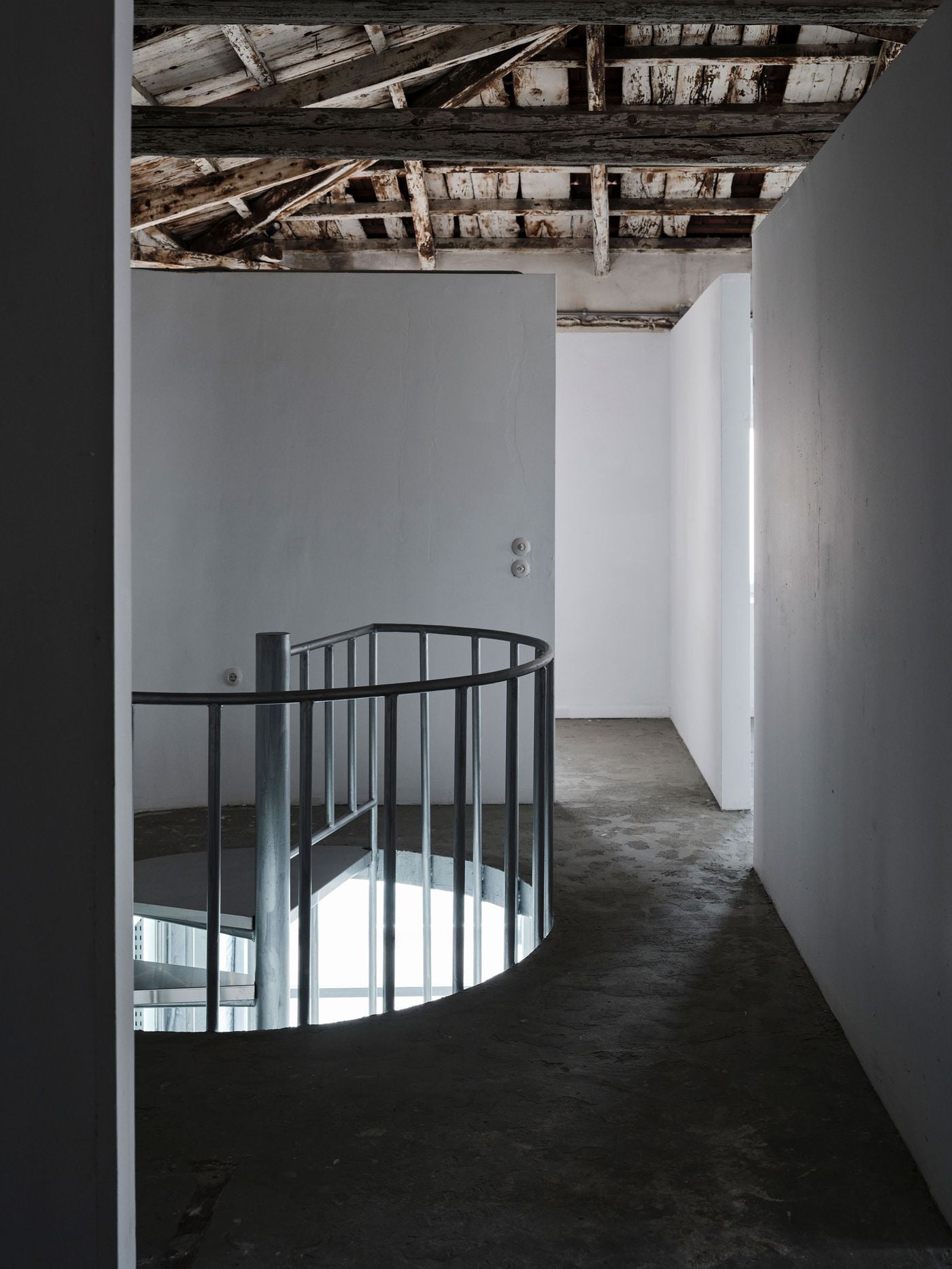 White partitions define the upper-level rooms without reaching the historic timber roof, allowing its weathered beams and boards to remain visually continuous overhead. A curved metal balustrade wraps around the stair opening, contrasting the rectilinear geometry of the partitions and guiding circulation between levels. White partitions define the upper-level rooms without reaching the historic timber roof, allowing its weathered beams and boards to remain visually continuous overhead. A curved metal balustrade wraps around the stair opening, contrasting the rectilinear geometry of the partitions and guiding circulation between levels.
