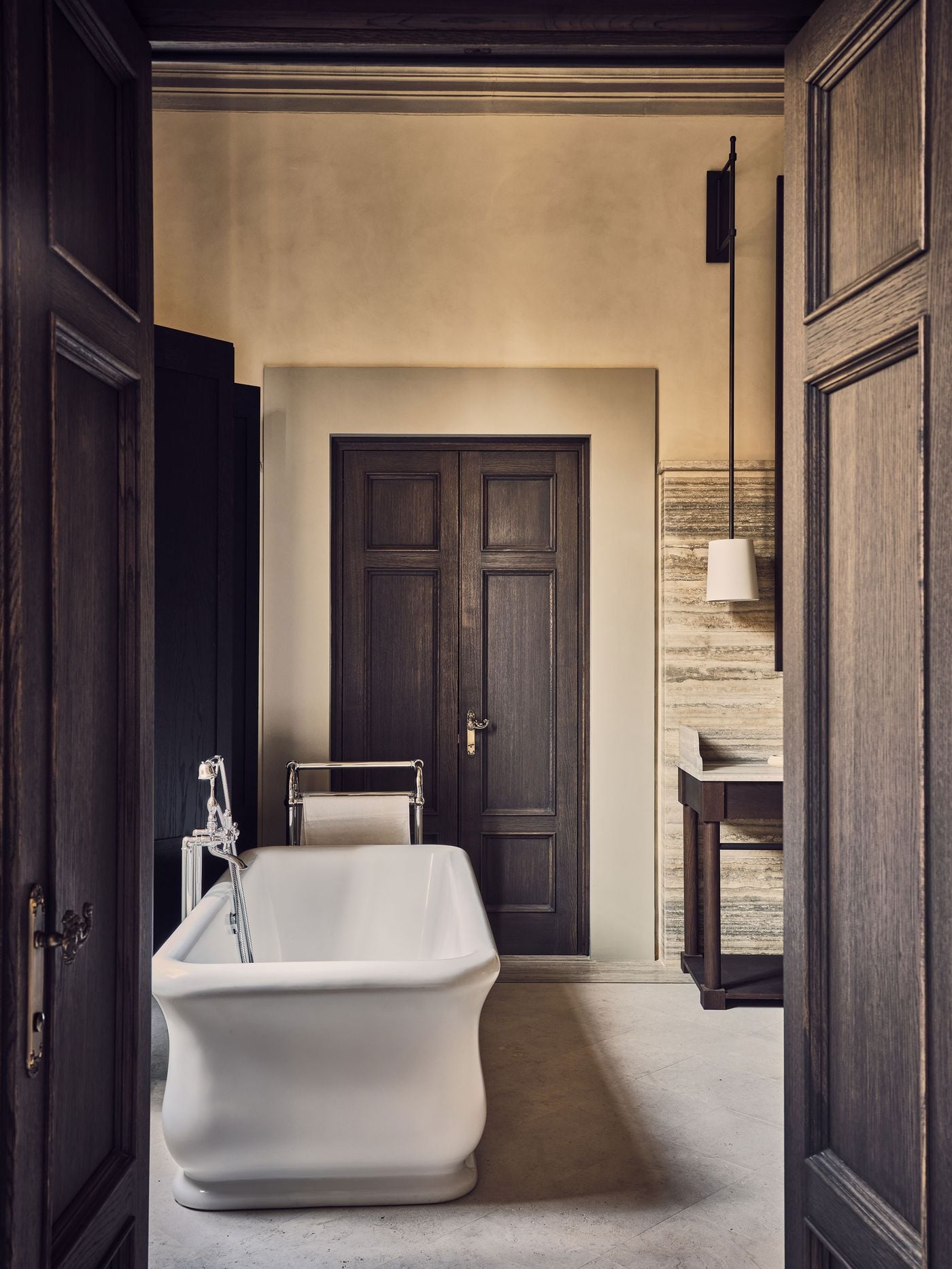 Framed by a pair of dark oak doors, a freestanding bathtub with softly curved sides takes centre stage in a bathroom at La Réserve Firenze. Polished nickel fittings, a banded travertine wainscot, a wall-mounted pendant and panelled double doors set a palette of cream, smoke and stone. Framed by a pair of dark oak doors, a freestanding bathtub with softly curved sides takes centre stage in a bathroom at La Réserve Firenze. Polished nickel fittings, a banded travertine wainscot, a wall-mounted pendant and panelled double doors set a palette of cream, smoke and stone.