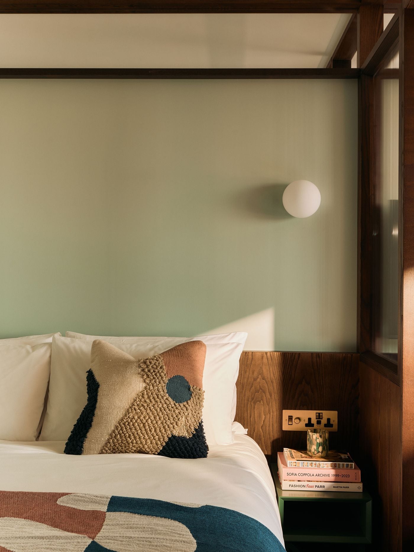 A restrained bedroom vignette pairs sage-green walls with walnut panelling, crisp white bedding and graphic textiles in rust, cream and blue. A simple globe wall light and compact bedside shelf reinforce the room’s quietly mid-century character, while angled sunlight gives the pared-back composition warmth and depth. A restrained bedroom vignette pairs sage-green walls with walnut panelling, crisp white bedding and graphic textiles in rust, cream and blue. A simple globe wall light and compact bedside shelf reinforce the room’s quietly mid-century character, while angled sunlight gives the pared-back composition warmth and depth.