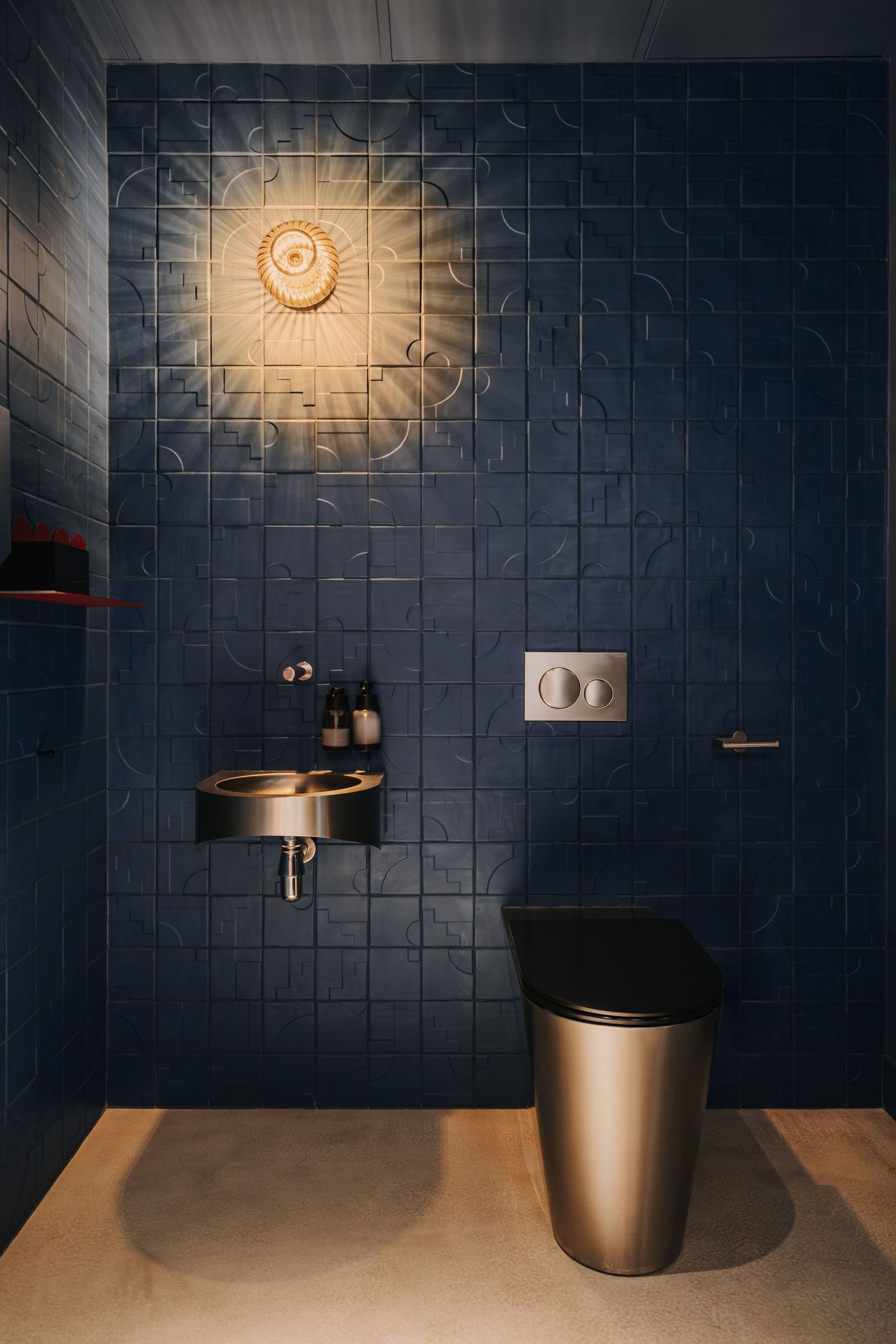 The bathroom at WIN wine bar, designed by Znamy się, is enveloped in deep indigo ceramic tiles embossed with abstract geometric motifs. A ribbed glass wall sconce casts dramatic radial shadows across the walls, while a compact stainless-steel washbasin and cylindrical metallic toilet introduce a sleek, utilitarian contrast. The bathroom at WIN wine bar, designed by Znamy się, is enveloped in deep indigo ceramic tiles embossed with abstract geometric motifs. A ribbed glass wall sconce casts dramatic radial shadows across the walls, while a compact stainless-steel washbasin and cylindrical metallic toilet introduce a sleek, utilitarian contrast.