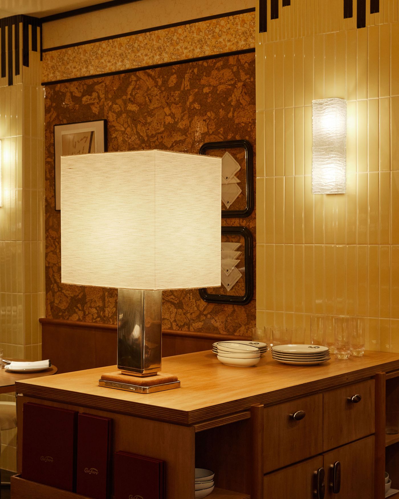 A wide, welcoming view of a dining area that skillfully combines Art Deco flair with mid-century design. A large, geometric lamp on a wooden counter anchors the space, while subtle lighting accentuates the interplay of glossy tiles and organic cork-like wall texture, creating a sophisticated and warm ambiance.