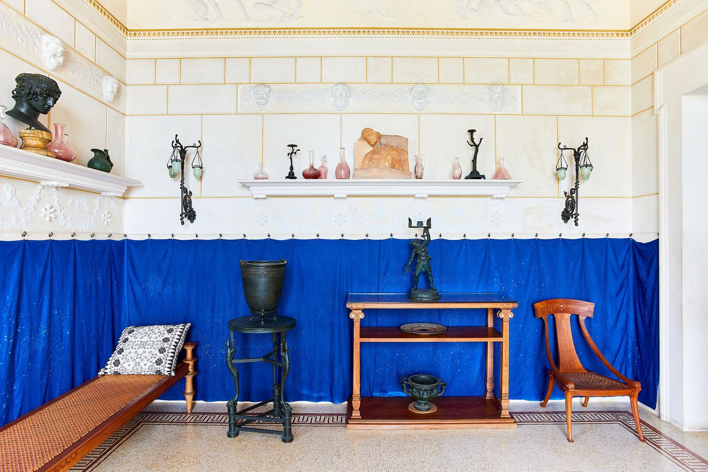 A wide shot of a room with a striking juxtaposition of styles. A lower half of vibrant blue curtain contrasts with the upper walls adorned with classical bas-reliefs and artifacts on floating shelves. A long wooden bench with a woven seat, a small table, and an iron stand with an urn create an eclectic composition, blending textures and materials. A wide shot of a room with a striking juxtaposition of styles. A lower half of vibrant blue curtain contrasts with the upper walls adorned with classical bas-reliefs and artifacts on floating shelves. A long wooden bench with a woven seat, a small table, and an iron stand with an urn create an eclectic composition, blending textures and materials.