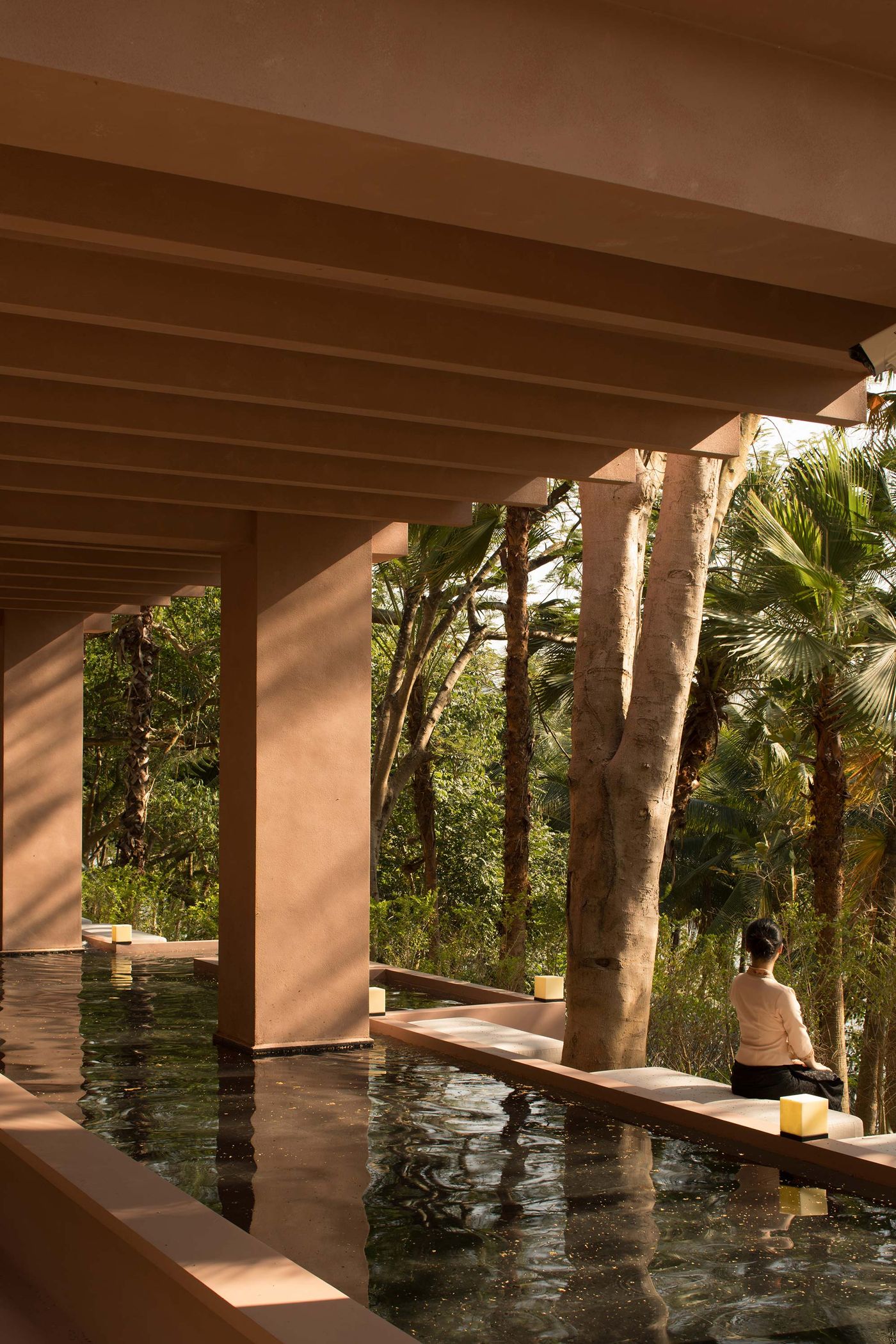A serene architectural space with a reflective water pool, framed by columns and surrounded by lush greenery, featuring a person sitting quietly. A serene architectural space with a reflective water pool, framed by columns and surrounded by lush greenery, featuring a person sitting quietly.