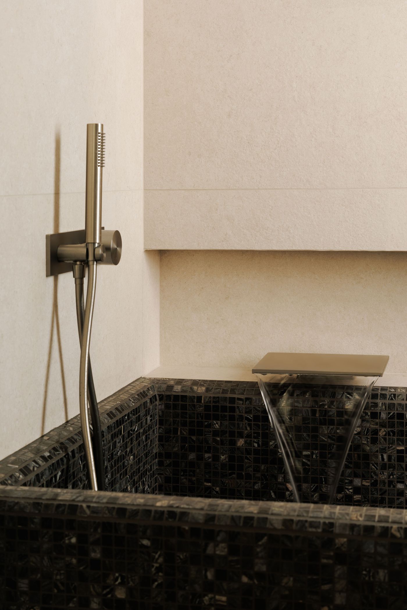 A close-up of a luxurious and artful bathing experience. A dark, richly textured mosaic-tiled bathtub features a unique, modern waterfall faucet that creates a mesmerizing cascade of water. A sleek, wall-mounted showerhead with a flexible hose in brushed metal completes the contemporary aesthetic, blending tactile richness with sophisticated functionality.