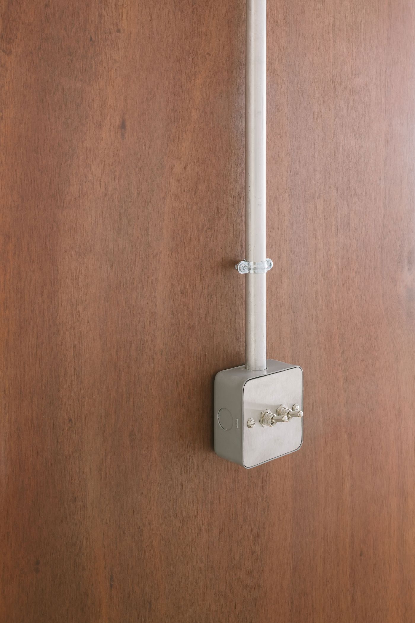 A gray electrical box with two switches and a vertical metal conduit, mounted on a brown wooden surface.
