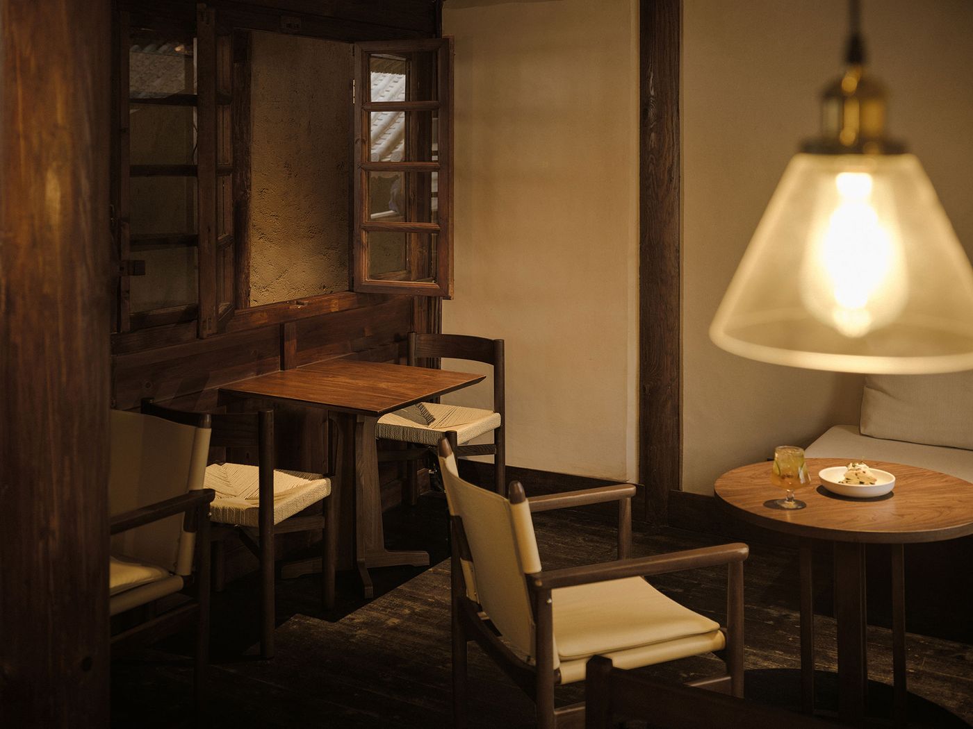 Cozy, warmly lit interior with wooden furniture, a small table, and chairs beside an open window, inviting relaxation.