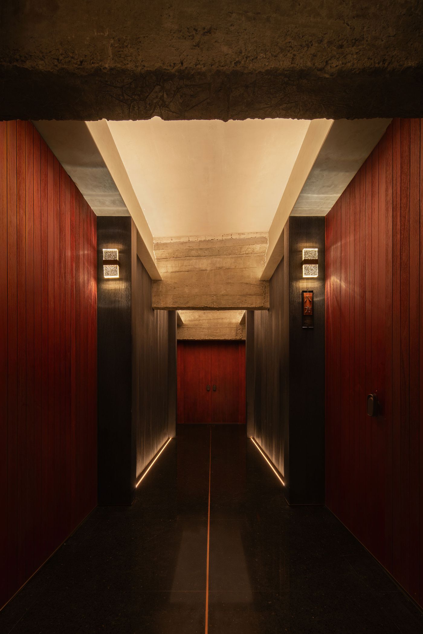 A modern, dimly lit hallway featuring rich wooden walls, soft lighting, and a striking dark floor leading to doors at the end. A modern, dimly lit hallway featuring rich wooden walls, soft lighting, and a striking dark floor leading to doors at the end.