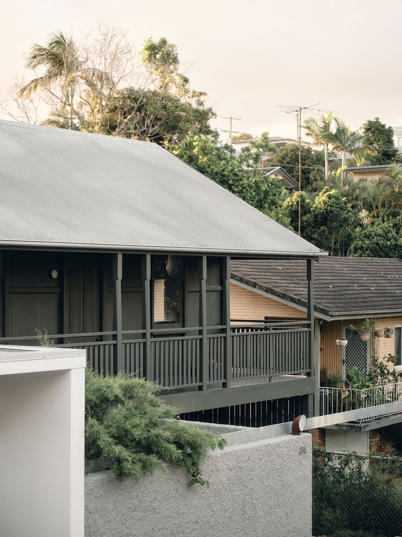A close-up view of a grey wooden house with a porch, surrounded by lush greenery and neighboring structures under a soft sky. A close-up view of a grey wooden house with a porch, surrounded by lush greenery and neighboring structures under a soft sky.