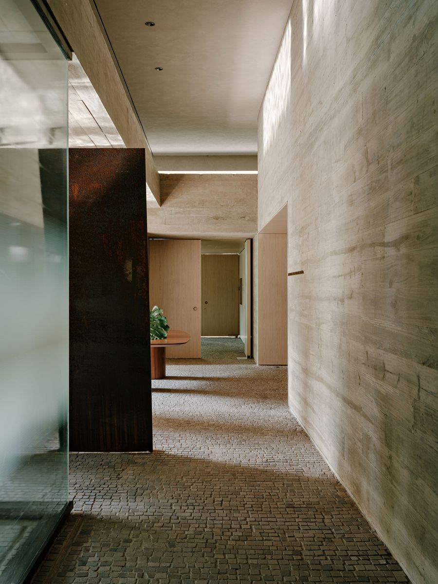 A dynamic perspective of this project's interior, where the rough texture of a concrete wall contrasts with the sleekness of a frosted glass partition and a rusted metal pivot door. The pathway of textured paving stones leads towards warm, light-filled rooms, embodying a refined industrial aesthetic with natural elements. A dynamic perspective of this project's interior, where the rough texture of a concrete wall contrasts with the sleekness of a frosted glass partition and a rusted metal pivot door. The pathway of textured paving stones leads towards warm, light-filled rooms, embodying a refined industrial aesthetic with natural elements.
