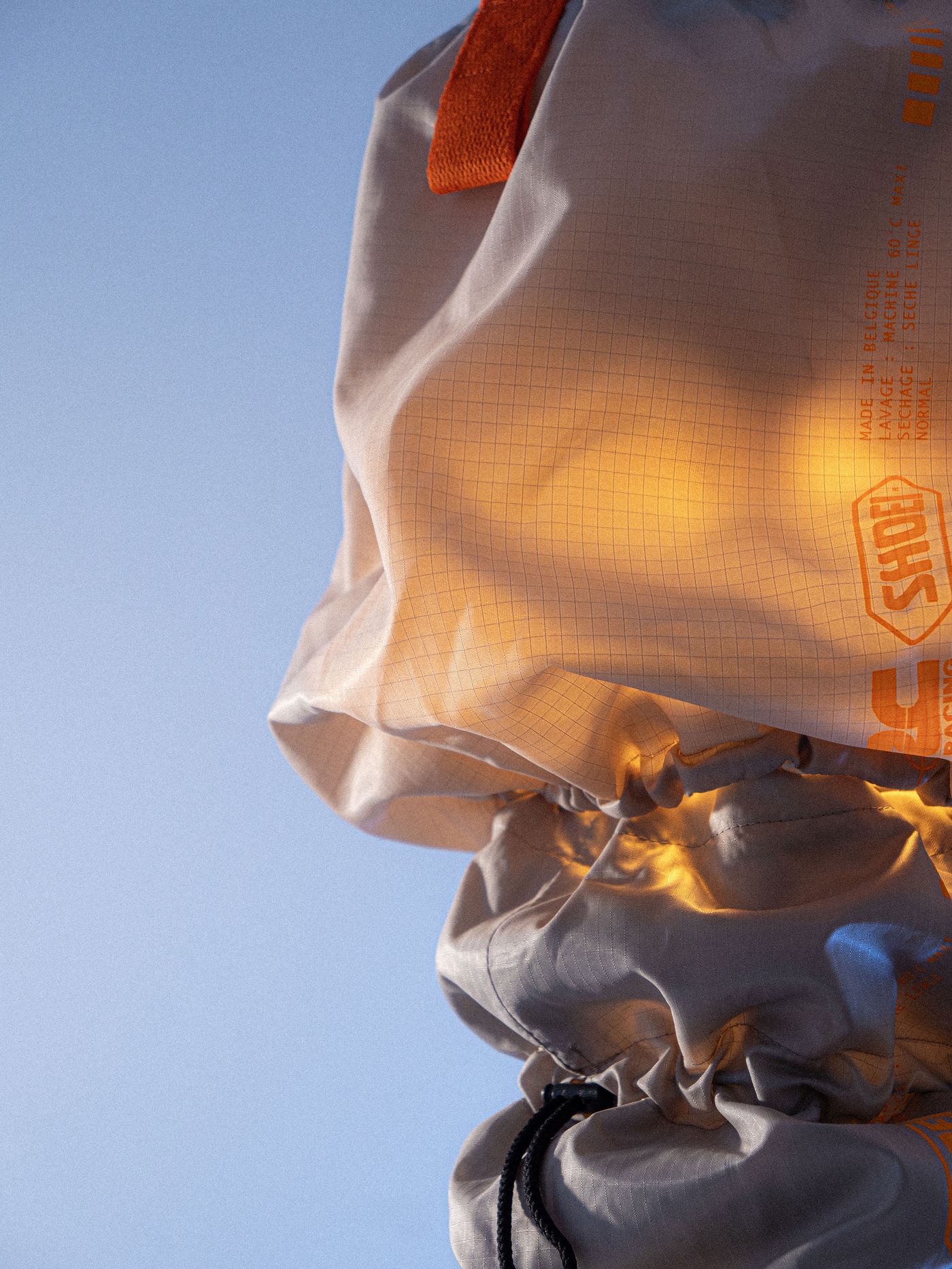 Close-up of a beige bag with an orange strap, featuring grid patterns and printed labels, against a clear blue sky. Close-up of a beige bag with an orange strap, featuring grid patterns and printed labels, against a clear blue sky.