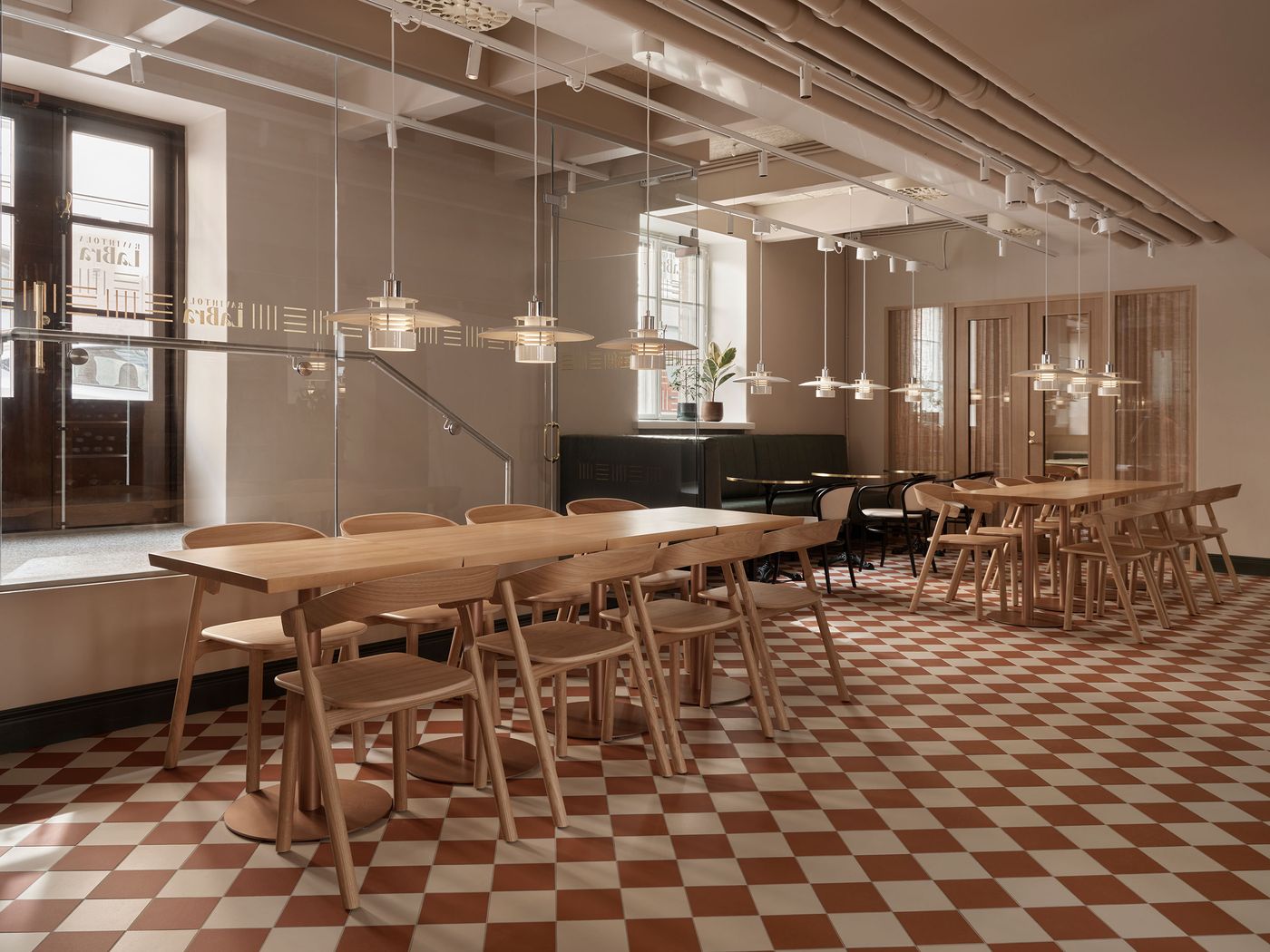 A spacious dining area at Konelabra, Helsinki by Fyra, featuring long wooden tables and minimalist chairs arranged on a classic red and white checkerboard tile floor. The industrial-chic space is warmly lit by multiple modern pendant lights suspended from exposed ceiling pipes. Large windows and a glass partition allow natural light to permeate, creating a bright, inviting atmosphere. A spacious dining area at Konelabra, Helsinki by Fyra, featuring long wooden tables and minimalist chairs arranged on a classic red and white checkerboard tile floor. The industrial-chic space is warmly lit by multiple modern pendant lights suspended from exposed ceiling pipes. Large windows and a glass partition allow natural light to permeate, creating a bright, inviting atmosphere.