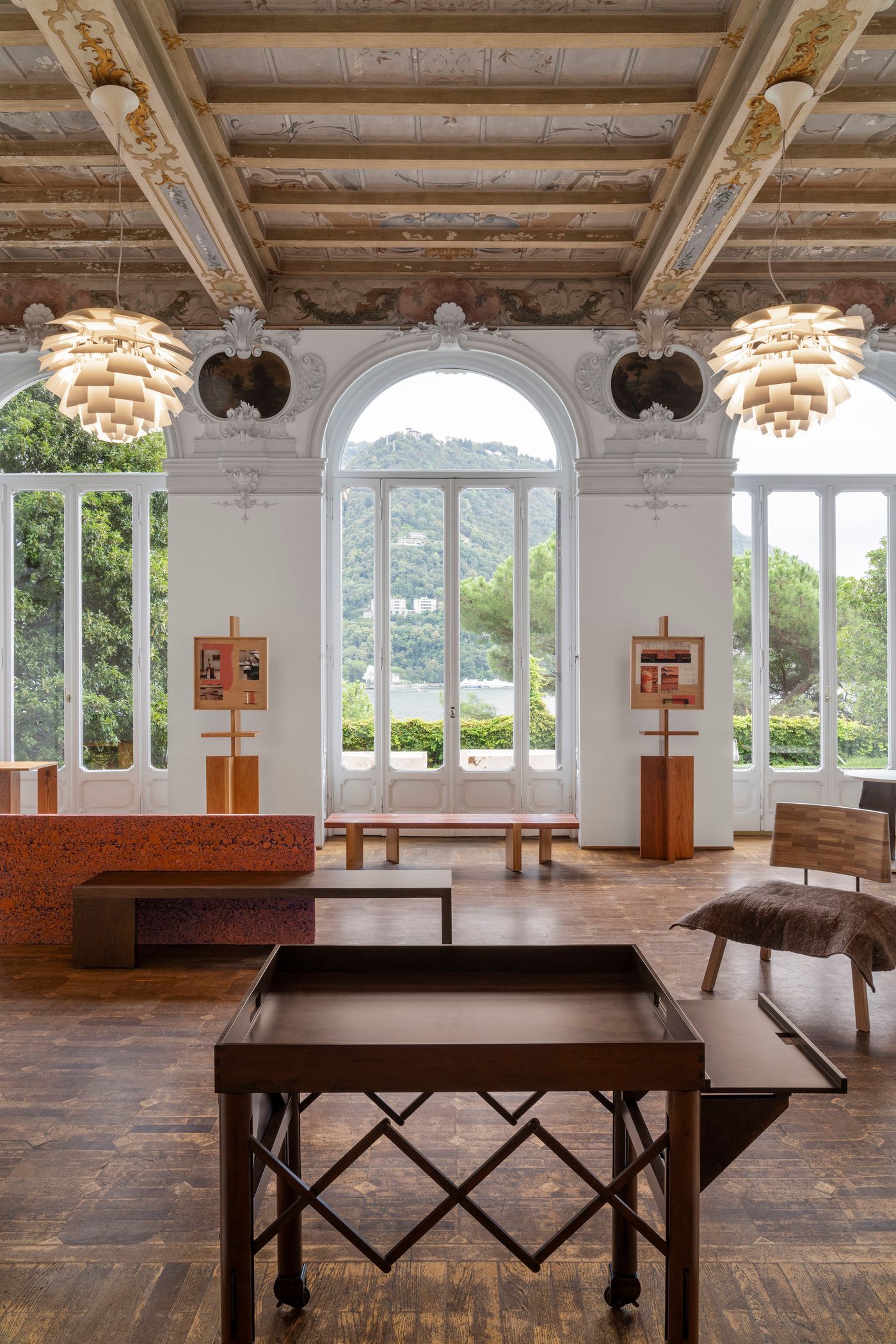 This grand room, with ornate plasterwork on the ceiling and large arched windows overlooking a lake and hills, displays a curated selection of contemporary furniture. The wooden floor is a rich, textured surface. Furniture pieces range from a dark wooden tray table with crisscross legs to a low bench with a textured red cushion, and a wooden chair draped with a sheepskin throw. The overall atmosphere is sophisticated and minimalist, highlighting the design of each piece against the backdrop of classical architecture.