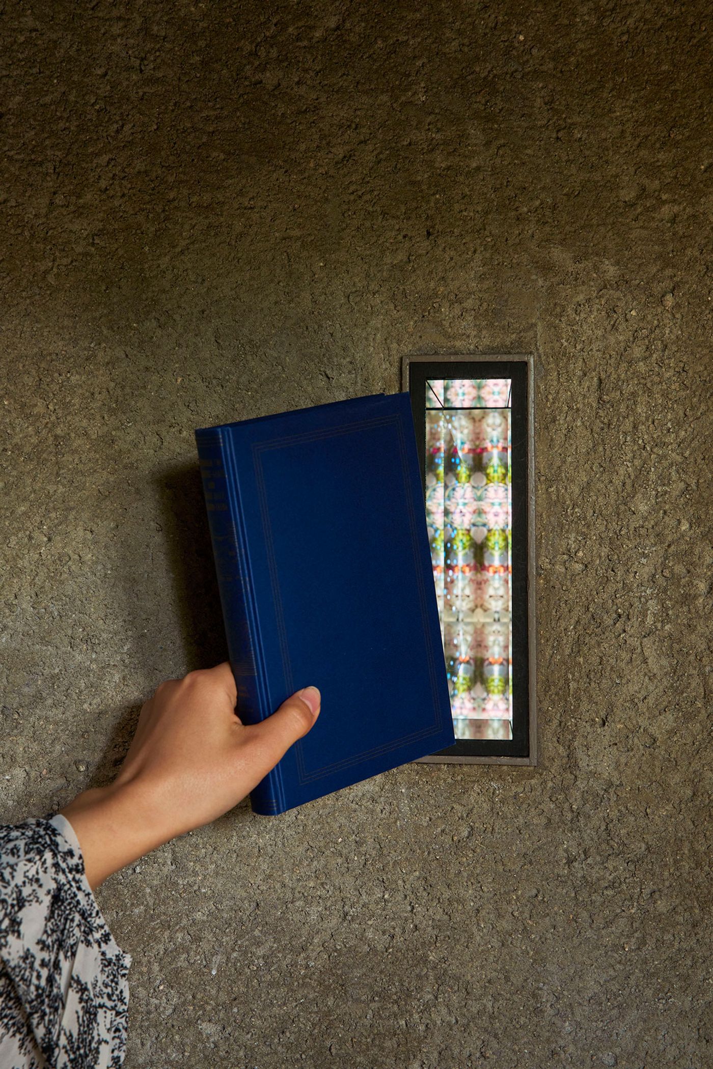 A hand holds a blue book near a narrow window revealing colorful, patterned glass against a textured concrete wall. A hand holds a blue book near a narrow window revealing colorful, patterned glass against a textured concrete wall.