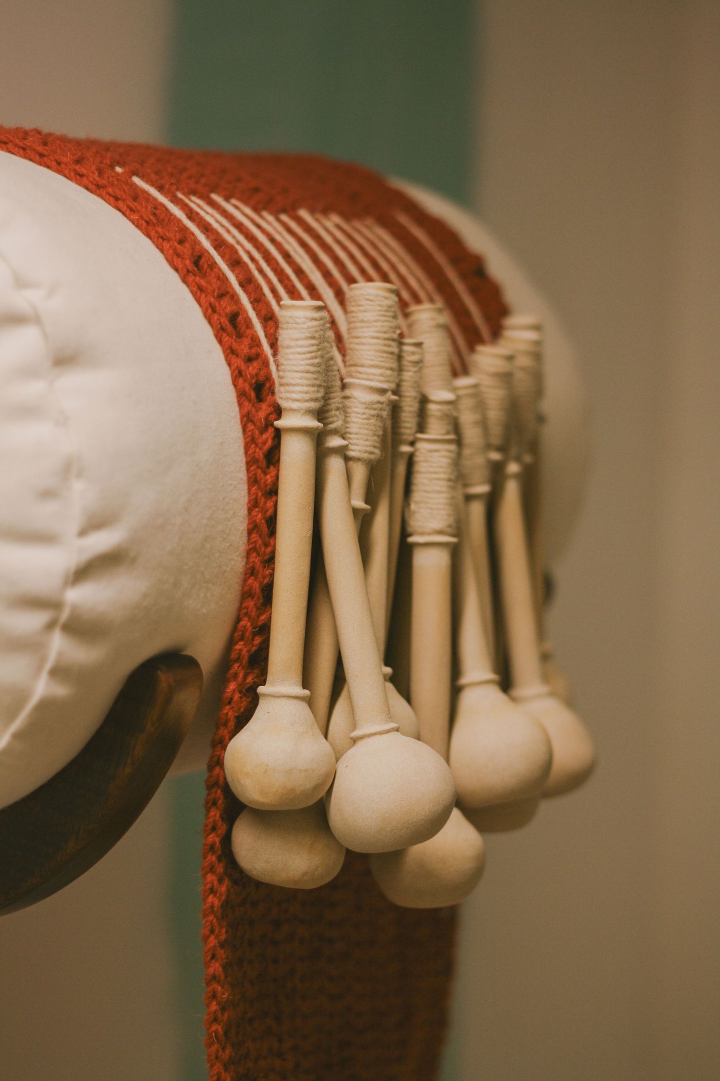 A close-up of an art piece from the "Crafting with AI" exhibition, featuring numerous light-toned, wooden, bobbin-like forms. They are intricately arranged against a vibrant reddish-orange knitted or woven fabric, draped over a soft white bolster. The composition highlights the delicate craftsmanship and the textural contrast between the smooth wood and the rich textile.