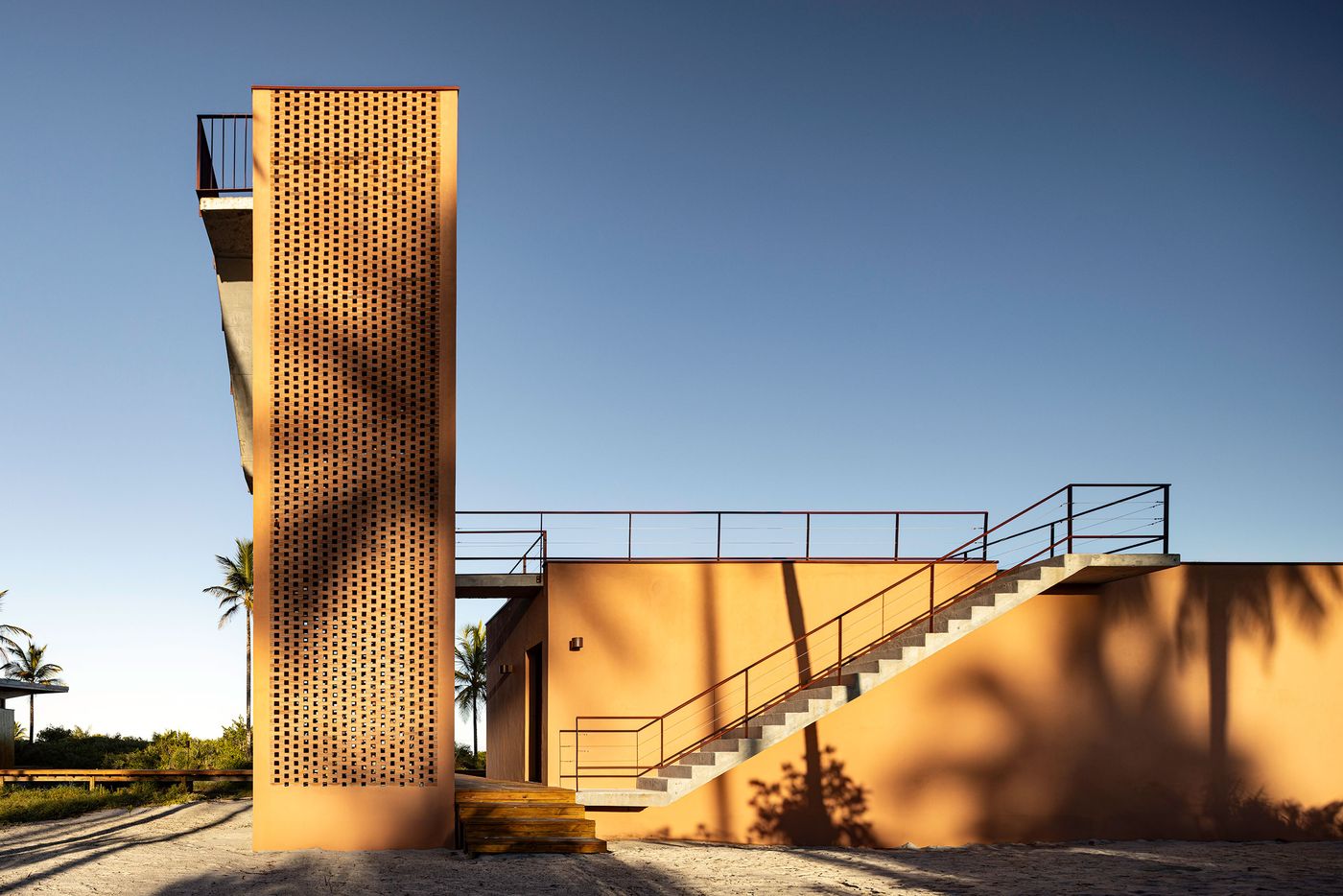 A striking, perforated brick tower in a warm peach hue stands beside a complementary building with an external staircase, all set against a clear blue sky. The geometric forms and textured materials create a modern, brutalist-inspired architectural statement in a tropical context. A striking, perforated brick tower in a warm peach hue stands beside a complementary building with an external staircase, all set against a clear blue sky. The geometric forms and textured materials create a modern, brutalist-inspired architectural statement in a tropical context.