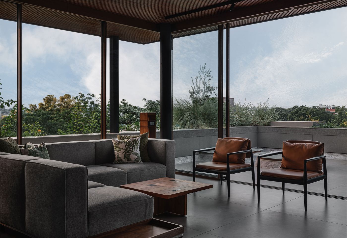 A contemporary living room with a dark grey sectional sofa and two tan leather armchairs, arranged to face expansive floor-to-ceiling windows. These windows offer a panoramic view of the natural landscape and sky. A slatted wooden ceiling and a simple wooden coffee table add warmth, creating a harmonious blend of indoor comfort and outdoor connection.