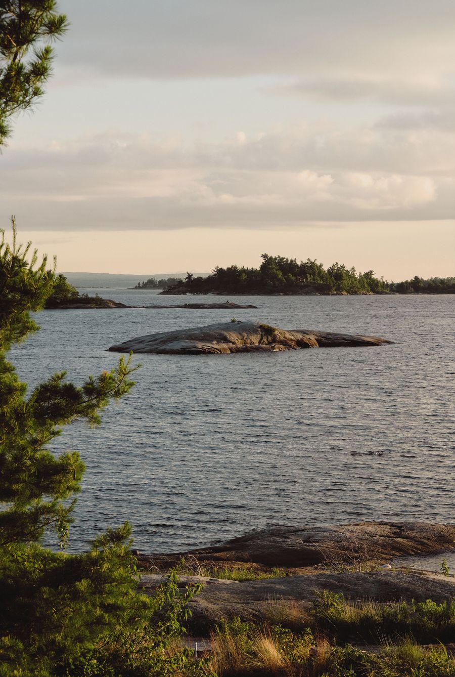 A serene lakeside view featuring rocky shores and island silhouettes under a soft, cloudy sky, framed by lush greenery. A serene lakeside view featuring rocky shores and island silhouettes under a soft, cloudy sky, framed by lush greenery.