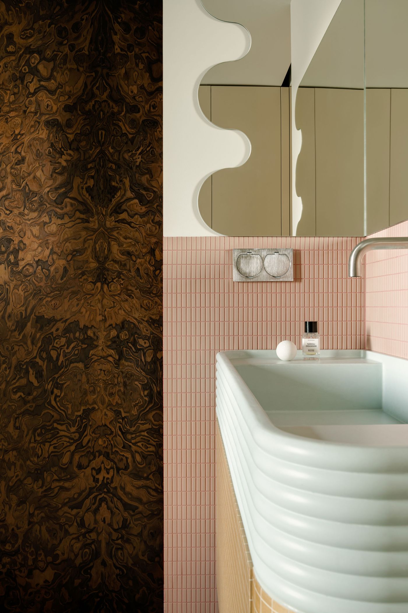 A close-up of the sculpted mirror and ribbed sink emphasizes the textured layering of materials: walnut burl veneer, glossy blush tiles, and a soft mint ceramic basin—revealing the designer’s flair for tactile contrast and visual rhythm.
