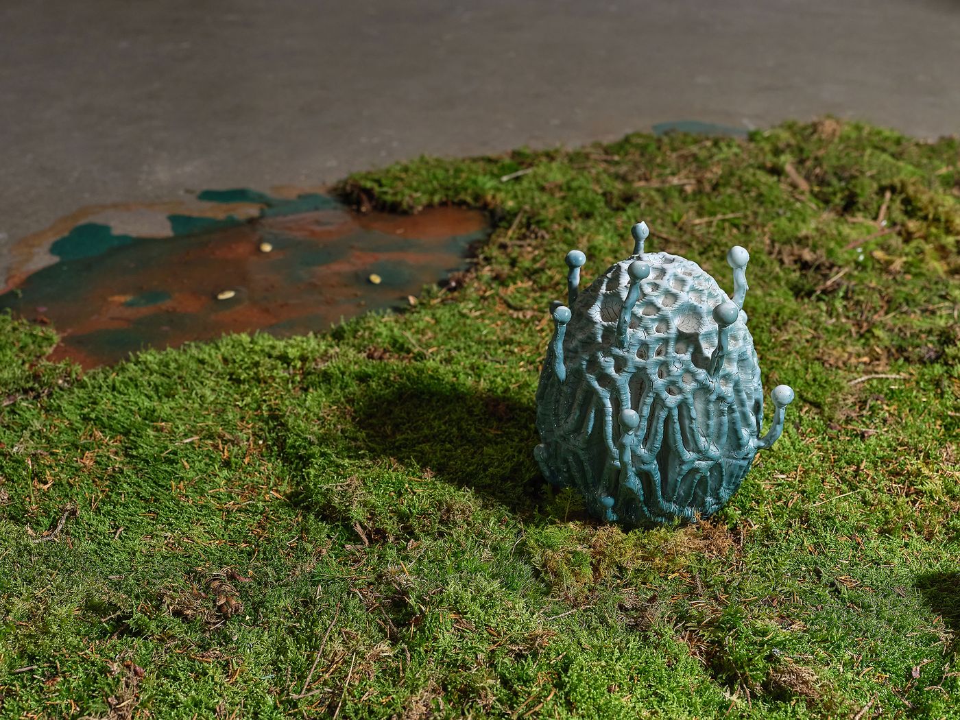 A textured, blue sculpture resembling a coral or alien egg sits on lush green moss, beside a small, rusty water puddle. A textured, blue sculpture resembling a coral or alien egg sits on lush green moss, beside a small, rusty water puddle.