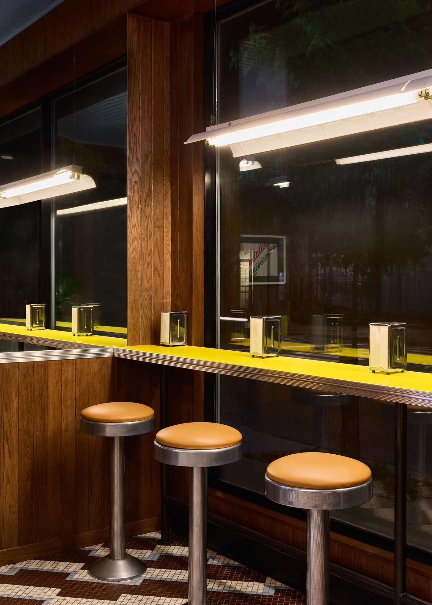 A view of the counter seating at Renzo Sandwich Shop, with three sleek, modern stools with tan seats set against a yellow counter. The backdrop of rich wood paneling and the geometric tile floor contribute to the establishment's retro-modern design. Large windows offer a view of the exterior, reflecting the interior lights.