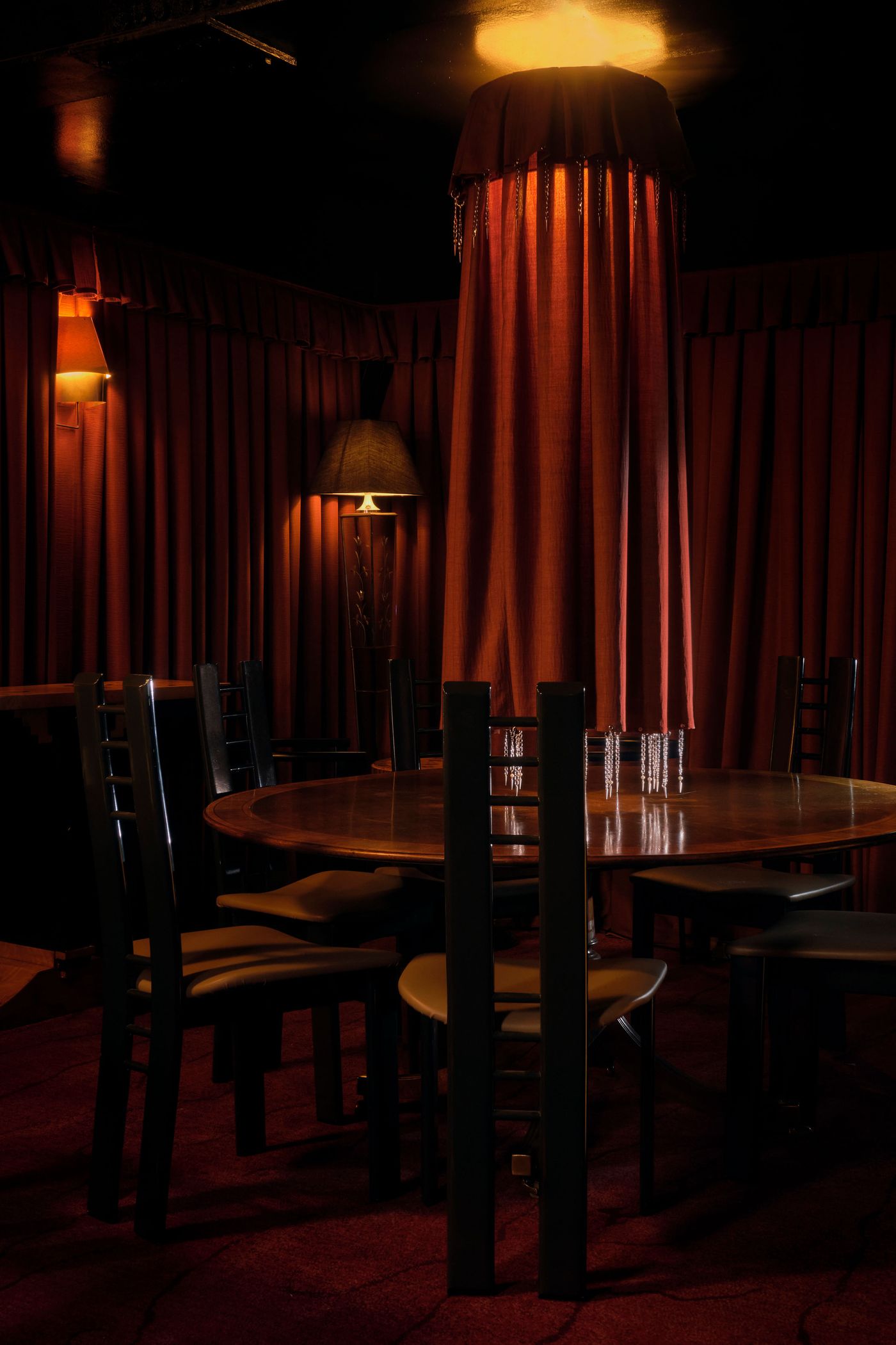 An intimate dining area in The Roses of Elagabalus, centered around a large, dark wooden round table with matching chairs. A unique cylindrical canopy of rich red fabric, suspended from the ceiling and adorned with hanging chains, defines the space, creating a sense of theatrical opulence and privacy, further enhanced by the surrounding red drapes and warm lighting. An intimate dining area in The Roses of Elagabalus, centered around a large, dark wooden round table with matching chairs. A unique cylindrical canopy of rich red fabric, suspended from the ceiling and adorned with hanging chains, defines the space, creating a sense of theatrical opulence and privacy, further enhanced by the surrounding red drapes and warm lighting.
