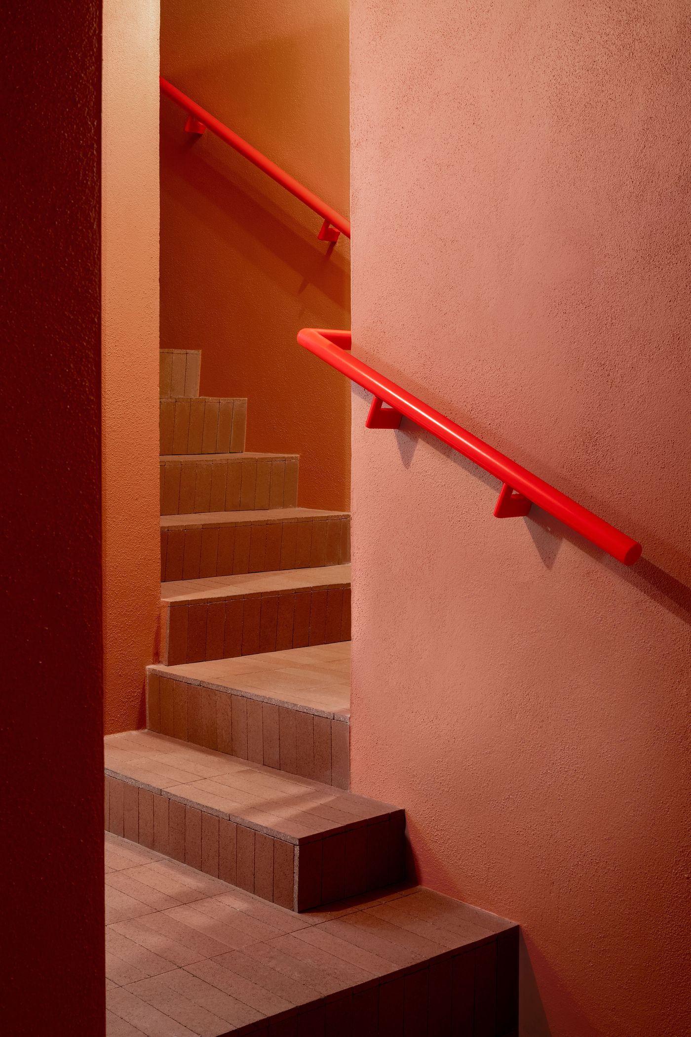 A striking view of a staircase within Santo Palato by Naessi Studio, characterized by warm terracotta-hued walls and contrasting bold red handrails. The steps, made of light-colored tile, ascend with a subtle glow, creating a dramatic and inviting transition between levels, emphasizing the restaurant's playful use of color and texture.
