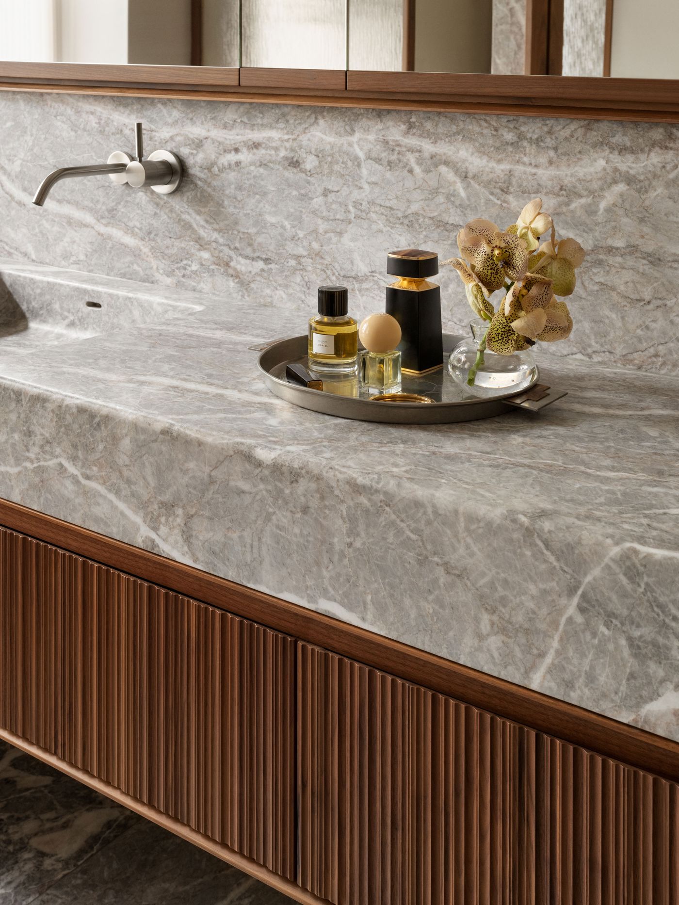 A luxurious bathroom sink with gray marble countertop, featuring a tray of elegant perfumes and a bouquet of orchids.