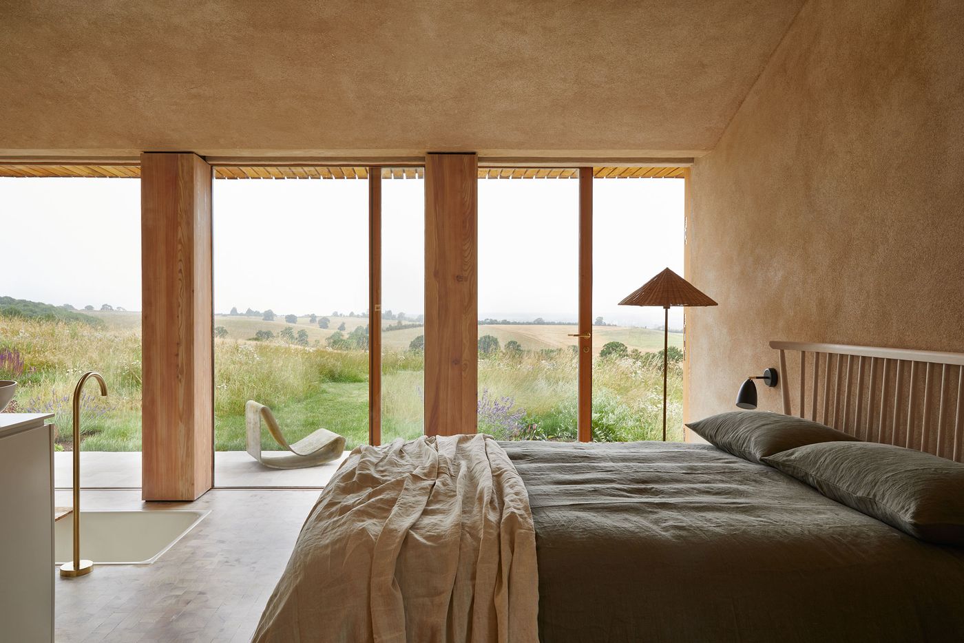 The bedroom in The Maker's Barn by Hutch Design offers a serene connection to the expansive rural landscape. Floor-to-ceiling windows, framed by warm timber, overlook fields of wild grasses and wildflowers. Inside, a dark-toned bed with simple bedding is complemented by a unique, sculptural floor lamp and a built-in basin with a brass faucet, blending minimalist design with rustic textures. The bedroom in The Maker's Barn by Hutch Design offers a serene connection to the expansive rural landscape. Floor-to-ceiling windows, framed by warm timber, overlook fields of wild grasses and wildflowers. Inside, a dark-toned bed with simple bedding is complemented by a unique, sculptural floor lamp and a built-in basin with a brass faucet, blending minimalist design with rustic textures.