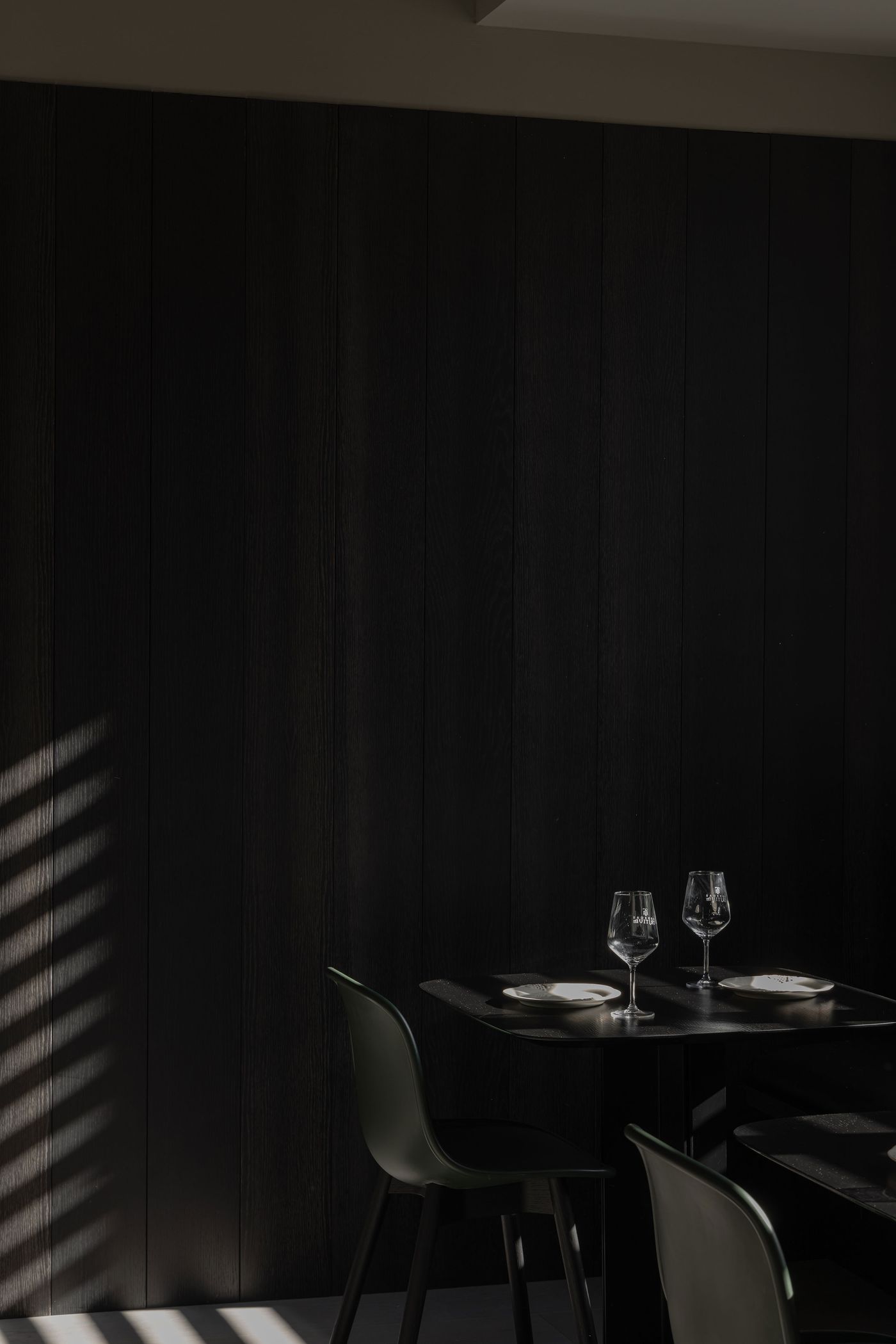 A dimly lit dining area features a black wooden wall, a sleek table set with two wine glasses and plates, and minimalist green chairs.