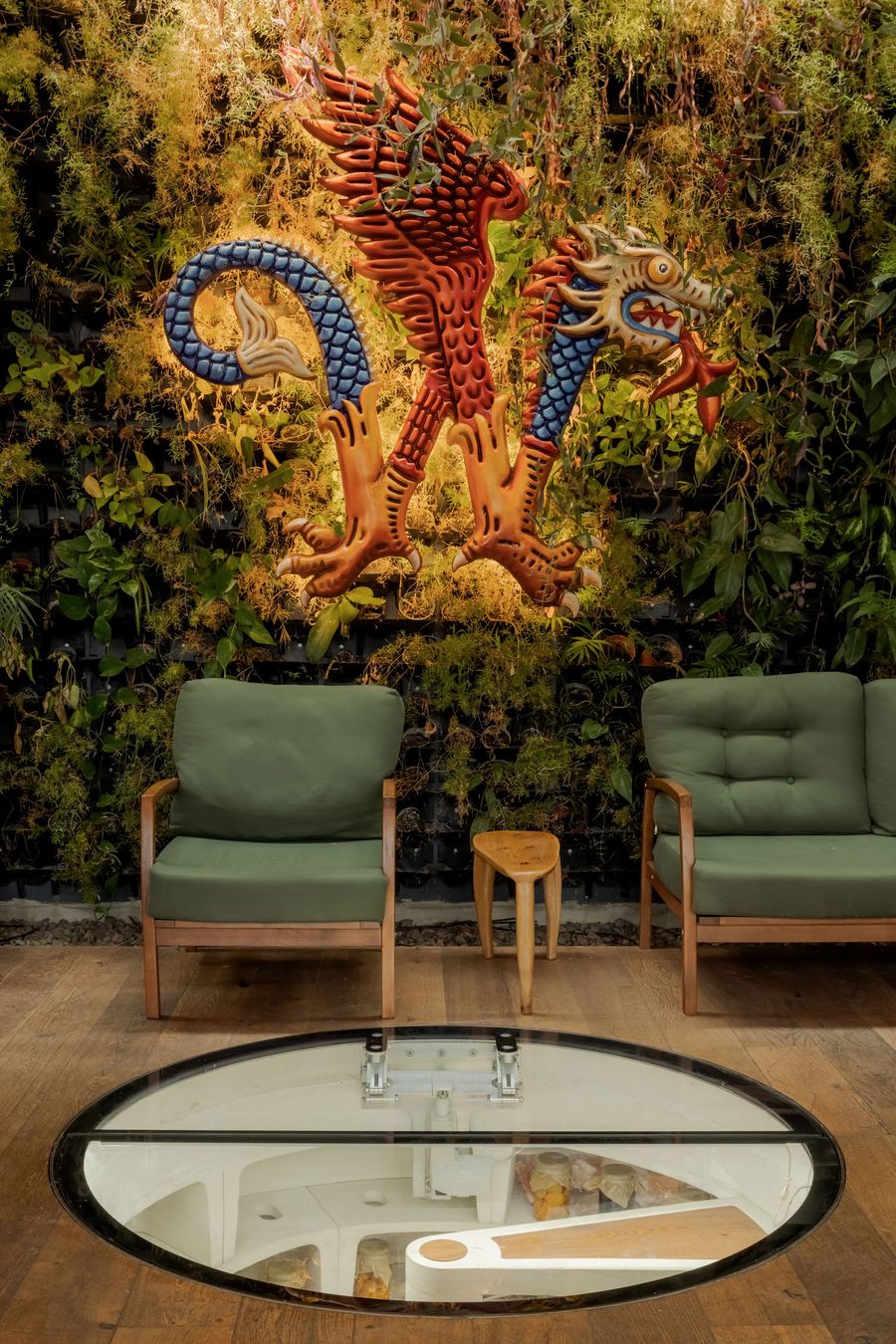 A vibrant wall art of a dragon and phoenix adorns a greenery-filled space with two green armchairs and a wooden stool. A vibrant wall art of a dragon and phoenix adorns a greenery-filled space with two green armchairs and a wooden stool.