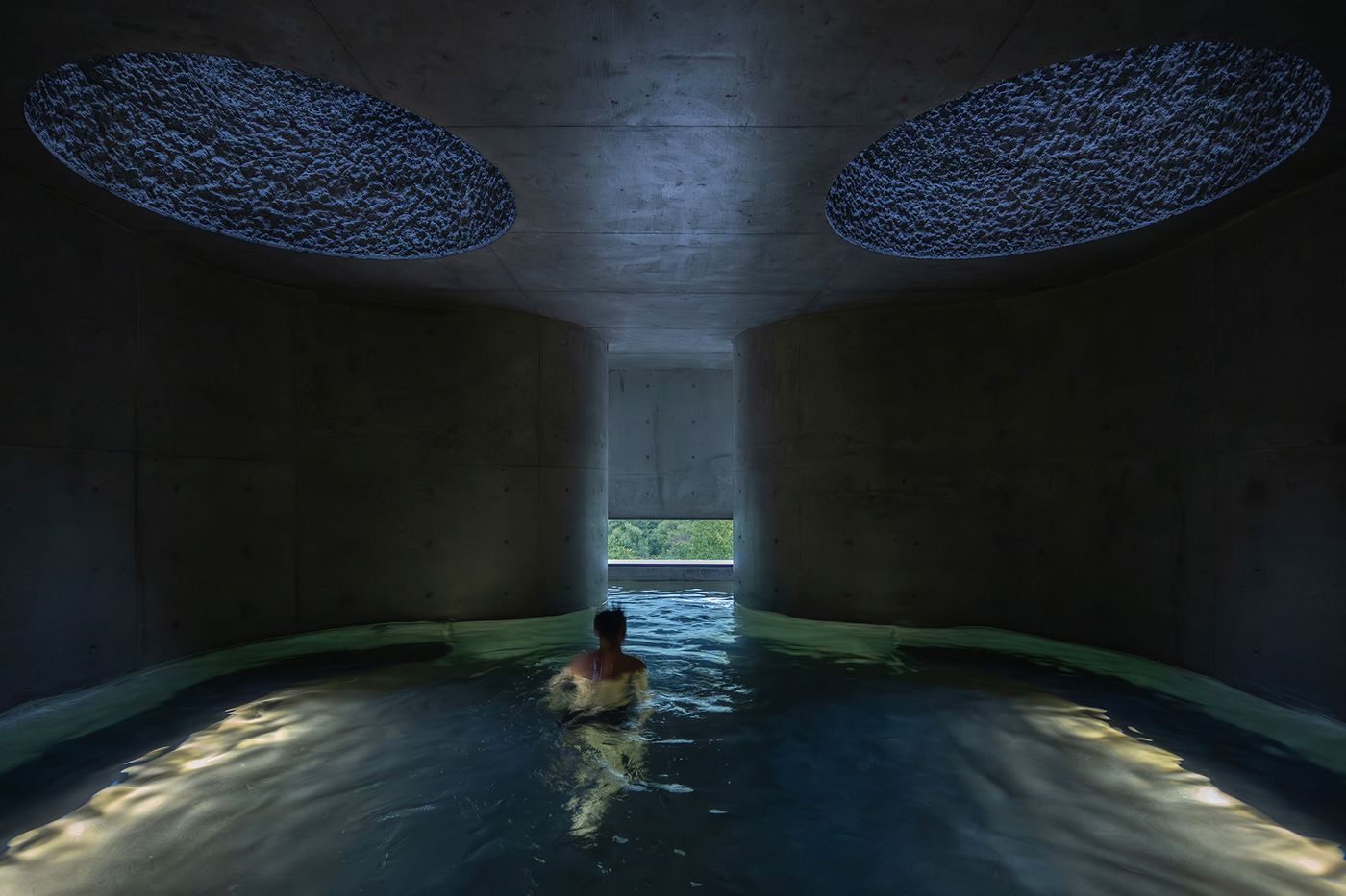 A person wades through a serene, dimly-lit pool beneath circular skylights, surrounded by smooth concrete walls. A person wades through a serene, dimly-lit pool beneath circular skylights, surrounded by smooth concrete walls.