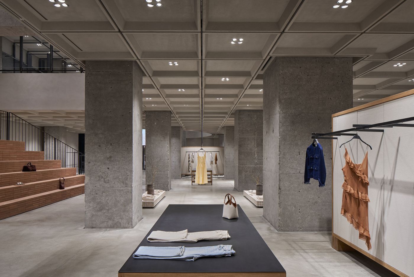 A modern clothing store interior featuring concrete pillars, minimalist displays, and a variety of garments on racks and tables.
