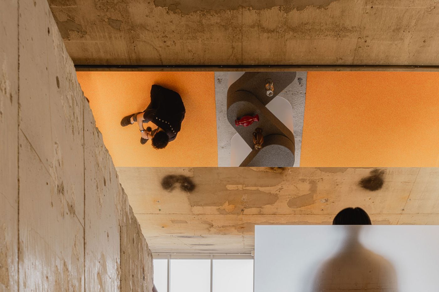 Andersson Bell Tokyo flagship by I IN: a mirrored ceiling reflects the upper floor from above, where a shopper in black crosses canary-yellow microcement flooring beside a serpentine display of stacked grey aggregate discs on frosted cylinders. Below, raw board-marked concrete and a softly blurred figure ground the vertiginous, disorienting composition.