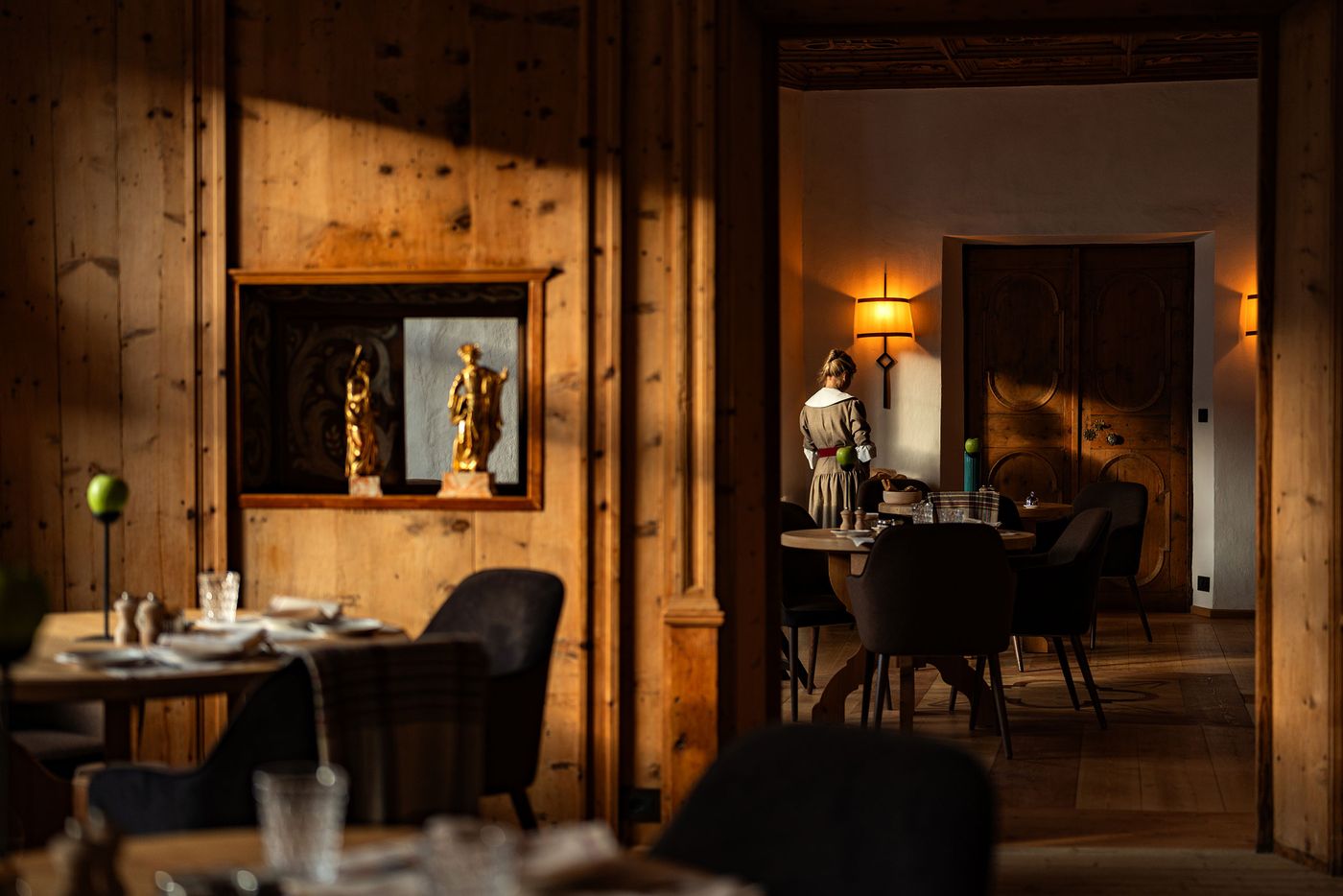 Late sunlight rakes across knotty pine panelling in Castel Badia's dining rooms, catching two gilded Baroque statuettes in a painted niche. Through an open doorway, a server in a traditionally styled dress lays a table beneath a glowing sconce, framed by carved double doors and a richly ornamented ceiling. Late sunlight rakes across knotty pine panelling in Castel Badia's dining rooms, catching two gilded Baroque statuettes in a painted niche. Through an open doorway, a server in a traditionally styled dress lays a table beneath a glowing sconce, framed by carved double doors and a richly ornamented ceiling.