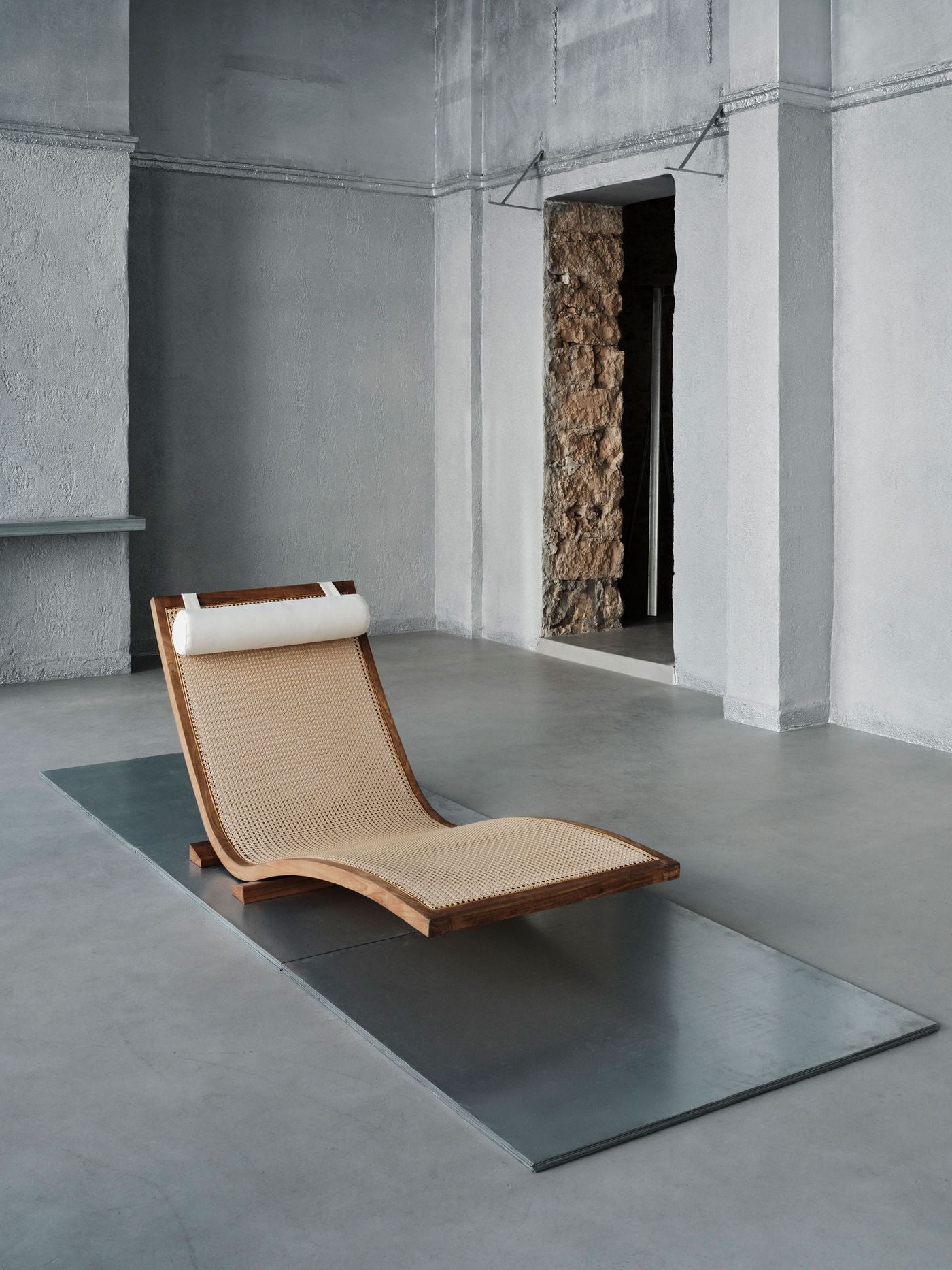 A sculptural timber-and-cane chaise rests on a rectangular metal platform within the monochromatic ground-floor interior. Its warm wood and woven surface stand out against silvery-grey walls and flooring, while a rough stone-lined doorway behind it reveals the historic masonry beneath MPNYC’s contemporary intervention. A sculptural timber-and-cane chaise rests on a rectangular metal platform within the monochromatic ground-floor interior. Its warm wood and woven surface stand out against silvery-grey walls and flooring, while a rough stone-lined doorway behind it reveals the historic masonry beneath MPNYC’s contemporary intervention.