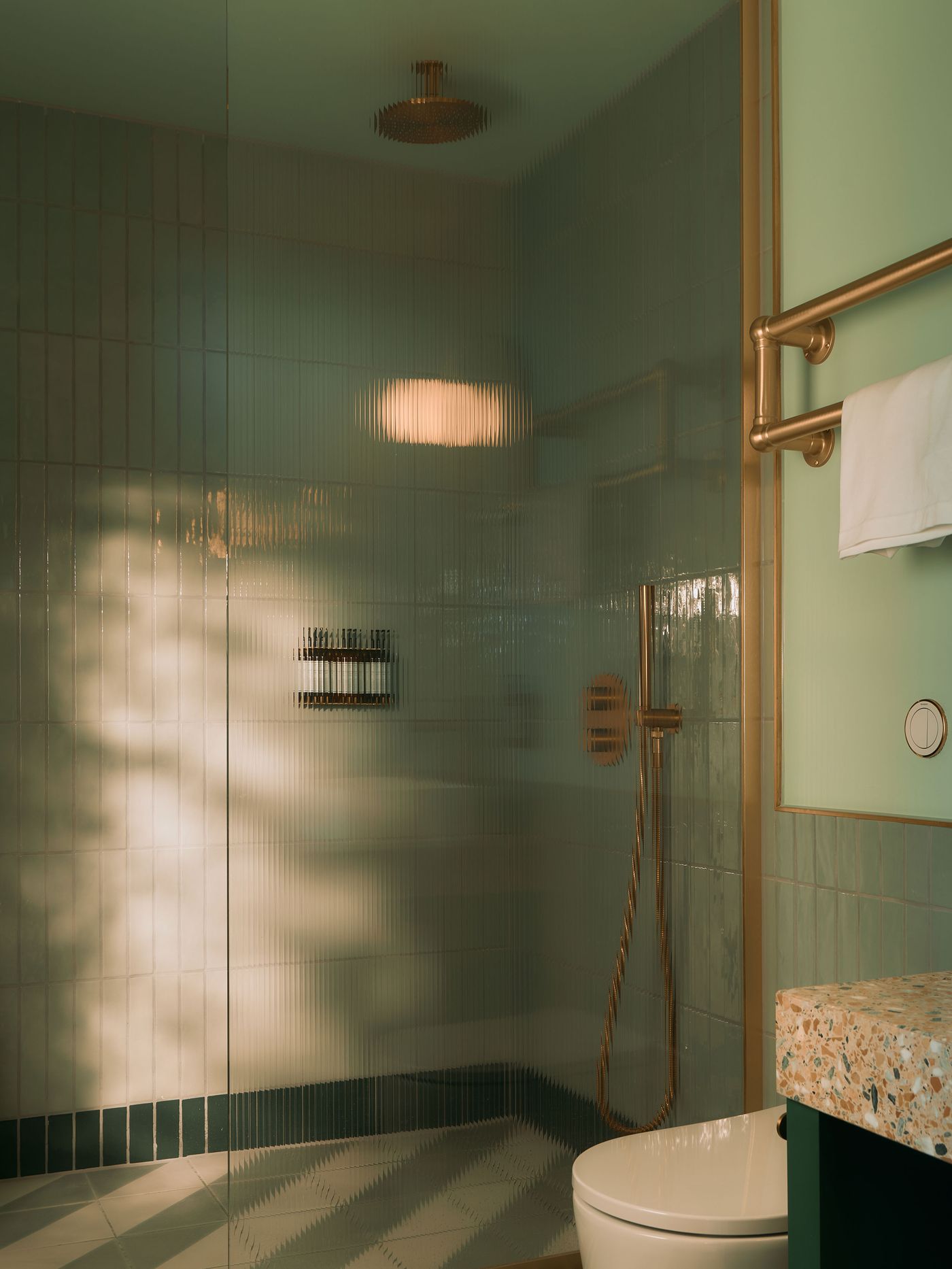 Reeded glass screens the guest-room shower, softly distorting the sage-green tiles and brass fittings beyond. Geometric floor tiles and a speckled terrazzo vanity continue the 1960s-inspired material palette, while filtered light gives the compact bathroom a muted, atmospheric quality rather than a purely functional feel. Reeded glass screens the guest-room shower, softly distorting the sage-green tiles and brass fittings beyond. Geometric floor tiles and a speckled terrazzo vanity continue the 1960s-inspired material palette, while filtered light gives the compact bathroom a muted, atmospheric quality rather than a purely functional feel.