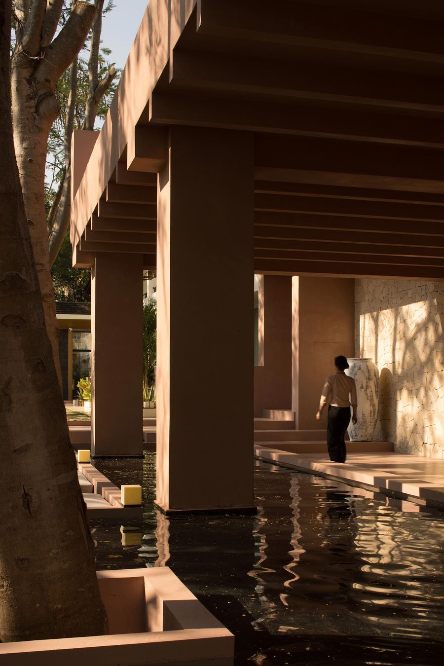 A person walks along a serene, water-lined path under a modern, peach-colored structure with geometric pillars and soft shadows. A person walks along a serene, water-lined path under a modern, peach-colored structure with geometric pillars and soft shadows.