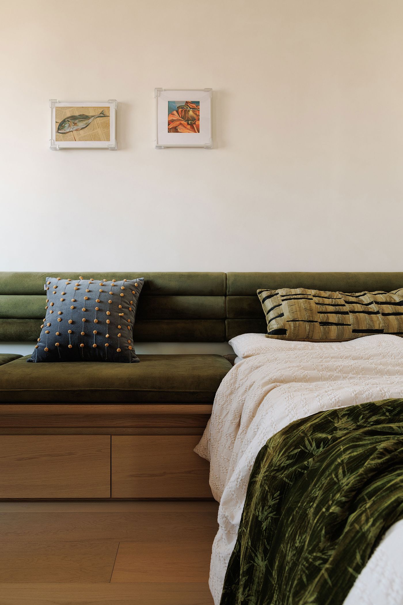 A versatile and inviting seating area, designed with a seamless transition between a daybed and a banquette. The olive green velvet upholstery with horizontal tufting is complemented by decorative pillows—one with dark blue and gold accents, another with textured earth tones. Two small framed artworks adorn the pale wall, adding a curated touch to this adaptable space.