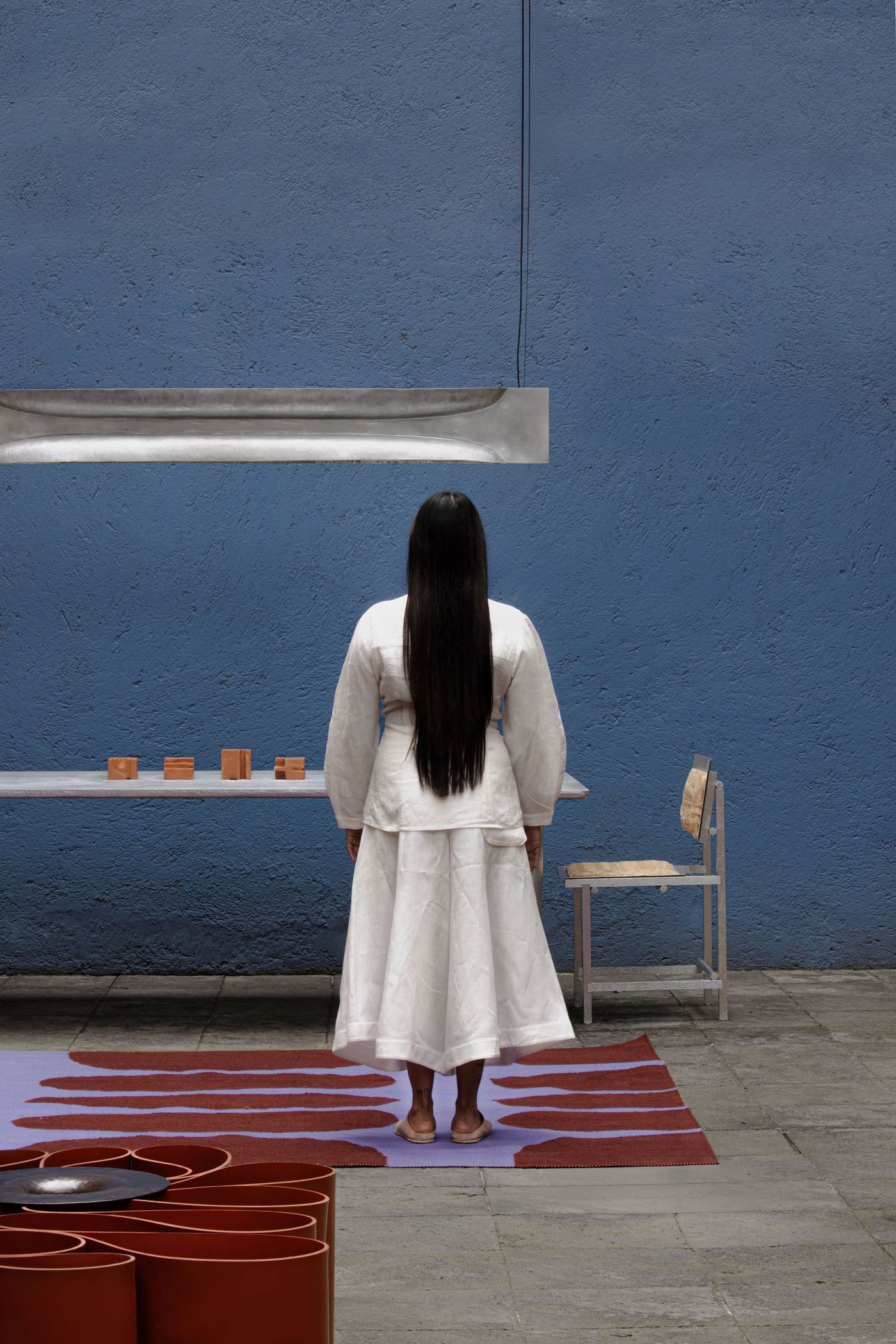 A woman in a white outfit stands in front of a blue wall, gazing at an art display on a table, with artistic decor around her.