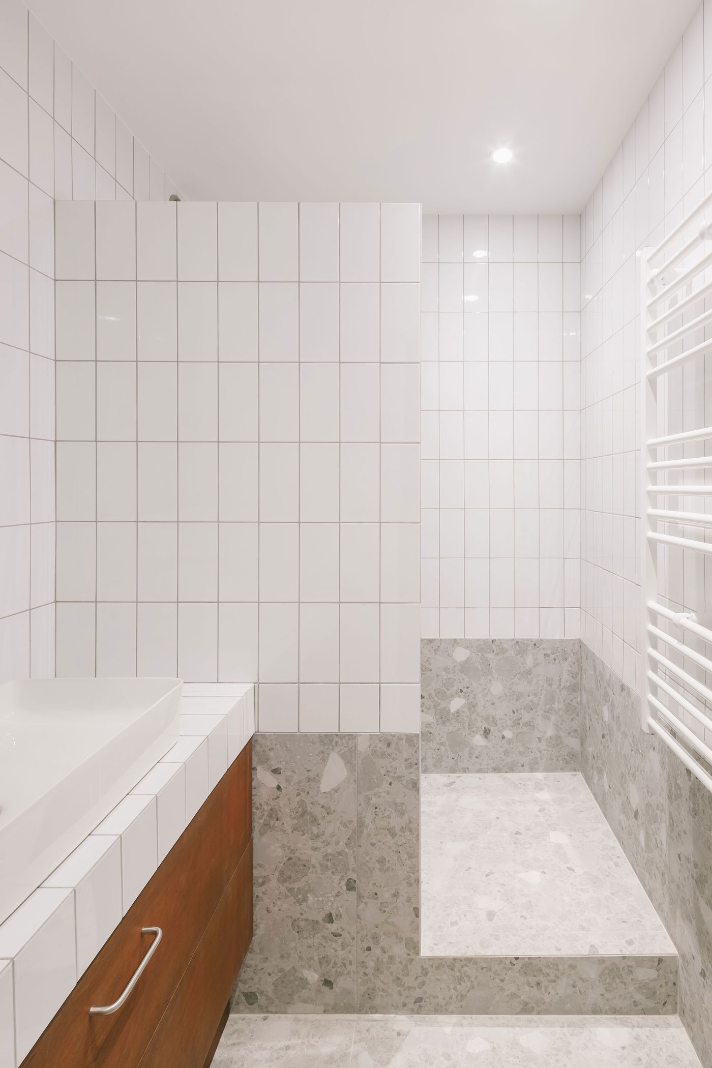 Modern bathroom featuring white tiled walls, a wooden cabinet, and a seamless shower area with textured stone flooring.