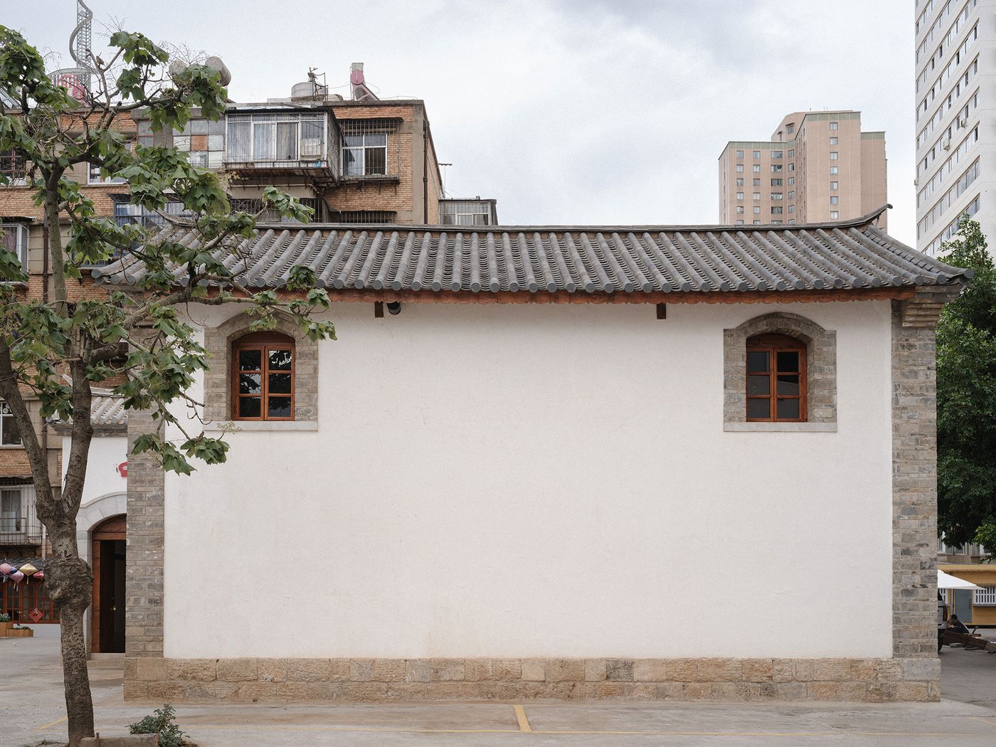 A simple white building with a stone base and traditional roof, surrounded by trees and contrasting modern high-rise buildings. A simple white building with a stone base and traditional roof, surrounded by trees and contrasting modern high-rise buildings.
