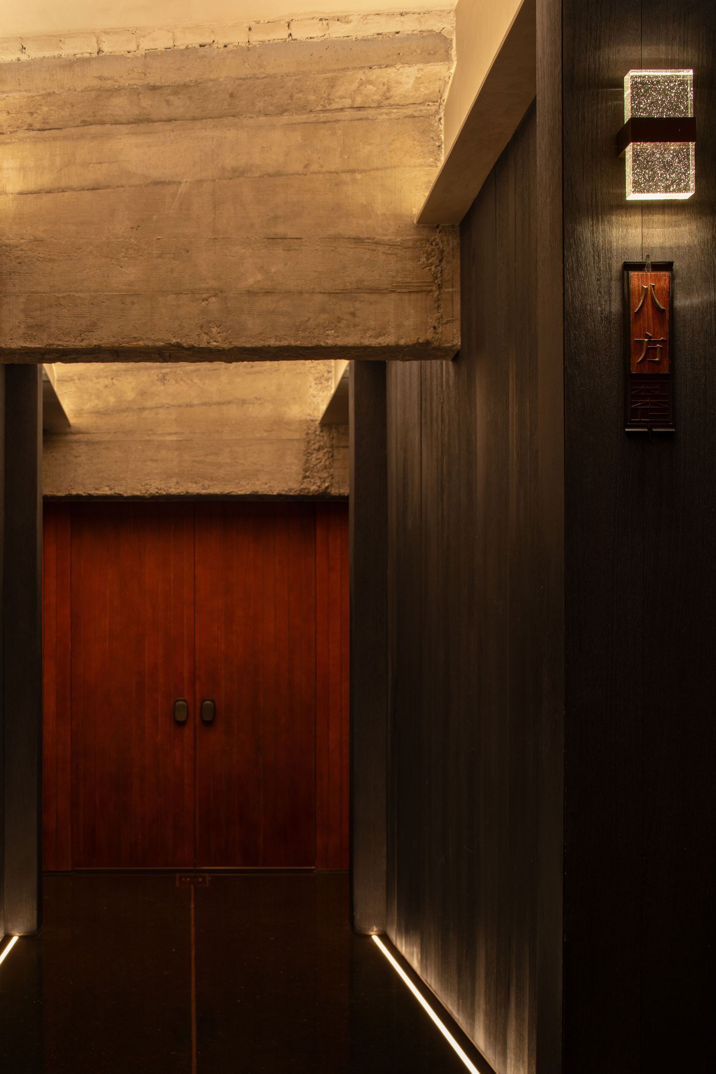Dimly lit hallway featuring textured concrete walls, dark wooden doors, and a glowing wall sconce with a wooden sign below. Dimly lit hallway featuring textured concrete walls, dark wooden doors, and a glowing wall sconce with a wooden sign below.