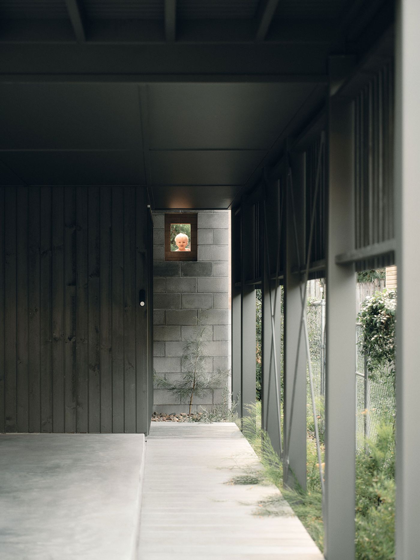 A minimalist corridor with dark wooden walls and a concrete floor leads to a garden outside, framed by large glass windows. A minimalist corridor with dark wooden walls and a concrete floor leads to a garden outside, framed by large glass windows.