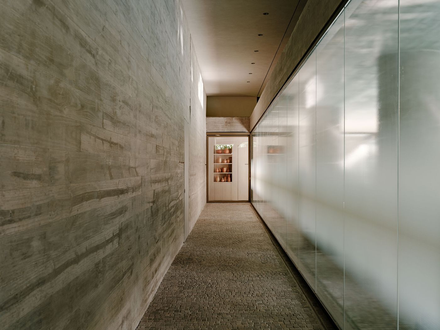 A long, artfully lit corridor, defined by a textured concrete wall on one side and a translucent glass wall on the other, creating a soft, diffused light. The interplay of materials and the subtle illumination emphasize the minimalist and serene atmosphere of the space. A long, artfully lit corridor, defined by a textured concrete wall on one side and a translucent glass wall on the other, creating a soft, diffused light. The interplay of materials and the subtle illumination emphasize the minimalist and serene atmosphere of the space.