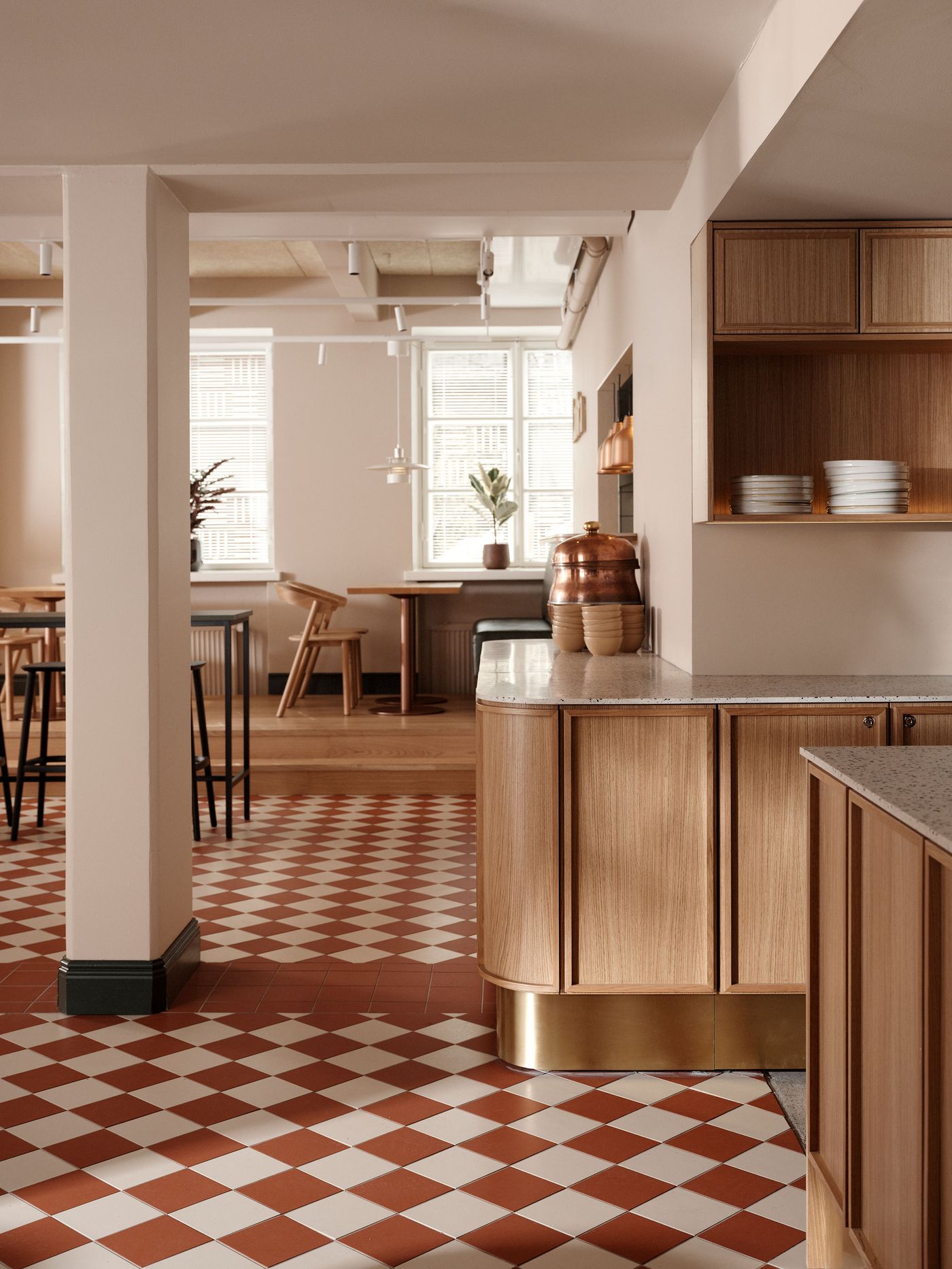 A charming dining and bar area at Konelabra, Helsinki by Fyra, distinguished by a classic red and white checkerboard tile floor. Curved, light wood cabinetry with brass accents defines the counter, adorned with copper pots. Soft, neutral walls and exposed ceilings blend industrial elements with warm, inviting Scandinavian aesthetics, complemented by natural light from large windows. A charming dining and bar area at Konelabra, Helsinki by Fyra, distinguished by a classic red and white checkerboard tile floor. Curved, light wood cabinetry with brass accents defines the counter, adorned with copper pots. Soft, neutral walls and exposed ceilings blend industrial elements with warm, inviting Scandinavian aesthetics, complemented by natural light from large windows.
