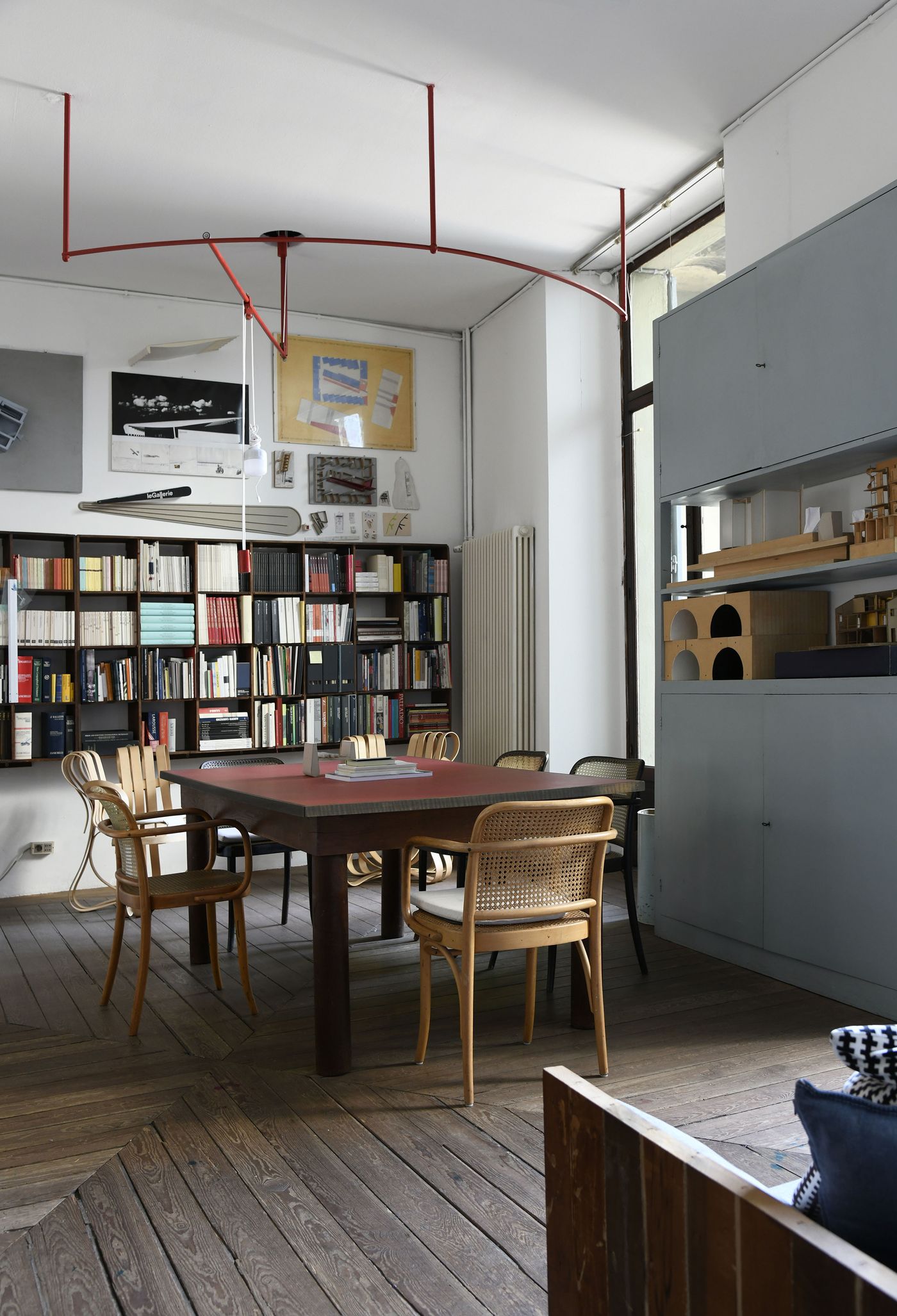 A room filled with books and art features a central table surrounded by wicker-backed chairs, illuminated by a unique red arc-shaped light fixture. A large grey cabinet with modular shelving units stands against the wall, displaying architectural models. The space has a lived-in, eclectic feel, with artworks and books contributing to a rich, intellectual atmosphere. The worn wooden floorboards add to the vintage charm.