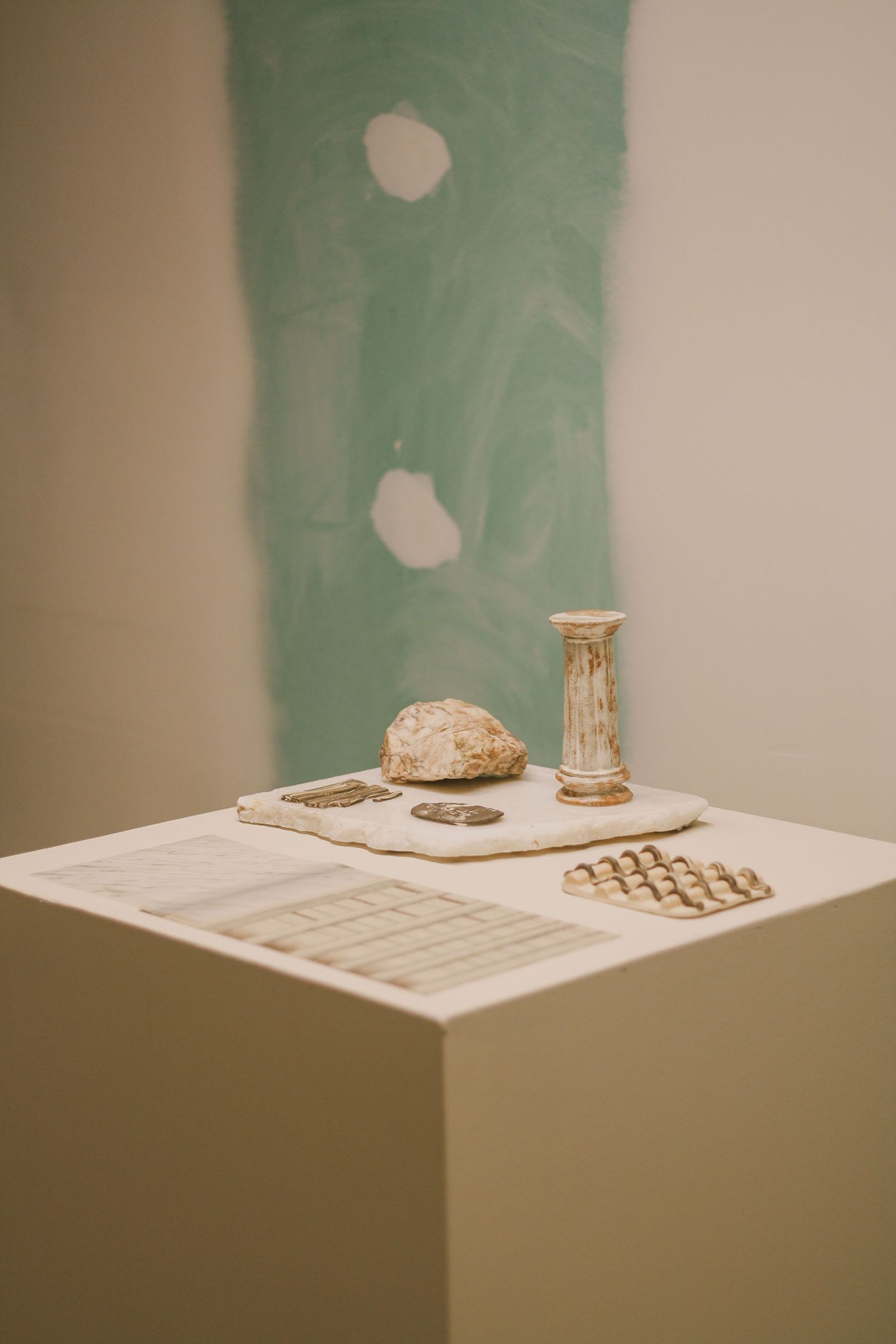 A contemplative still life from the "Crafting with AI" exhibition at Estrela 74, featuring a small stone column, a natural rock, and patterned flat objects on a white plinth. The background reveals a partially painted wall with visible plaster patches, hinting at an unfinished or deconstructed aesthetic. The composition emphasizes raw materials and an interplay between natural and crafted forms. A contemplative still life from the "Crafting with AI" exhibition at Estrela 74, featuring a small stone column, a natural rock, and patterned flat objects on a white plinth. The background reveals a partially painted wall with visible plaster patches, hinting at an unfinished or deconstructed aesthetic. The composition emphasizes raw materials and an interplay between natural and crafted forms.