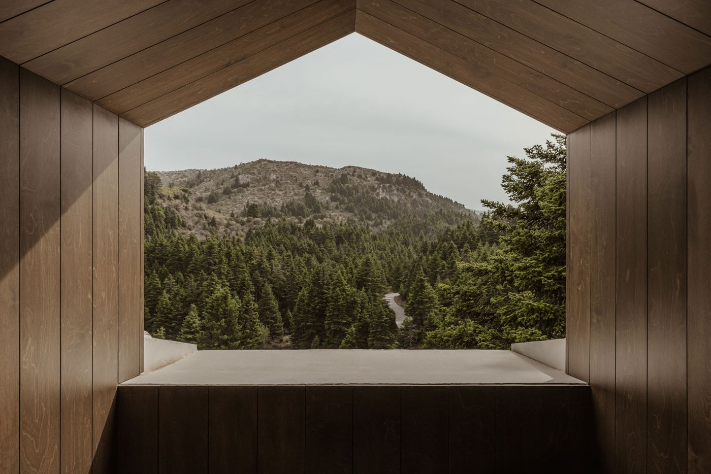 An architectural detail frames a captivating view of a mountainous landscape. The interior features warm wooden paneling on a sloped ceiling and walls, creating a cozy, almost meditative alcove. The large, unadorned opening perfectly captures the dense, verdant forest, emphasizing a deep connection to nature within a minimalist setting. An architectural detail frames a captivating view of a mountainous landscape. The interior features warm wooden paneling on a sloped ceiling and walls, creating a cozy, almost meditative alcove. The large, unadorned opening perfectly captures the dense, verdant forest, emphasizing a deep connection to nature within a minimalist setting.