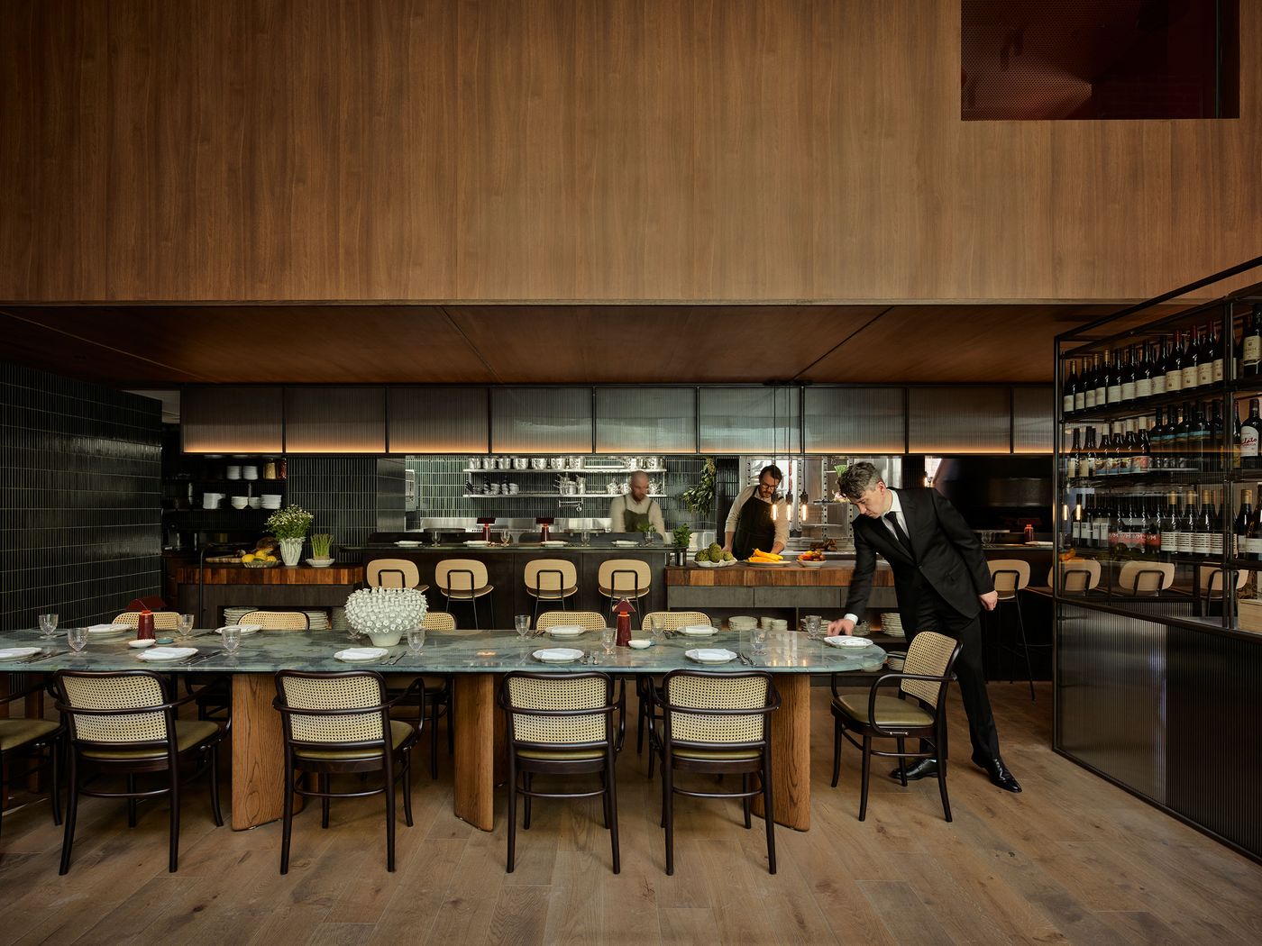 A sleek restaurant interior featuring a long dining table, elegant chairs, and a well-stocked wine display, with staff preparing food.