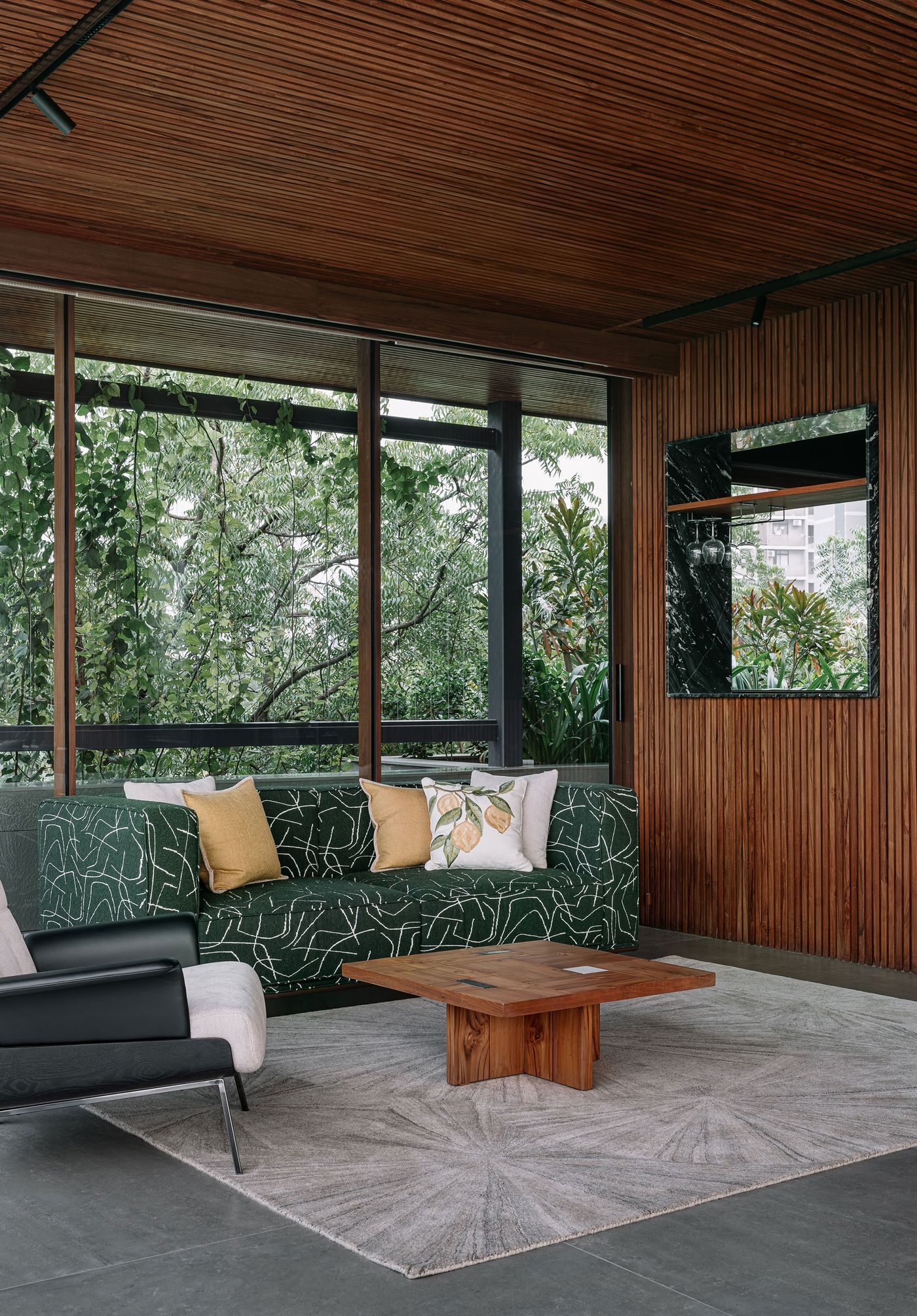 A sophisticated living space featuring a deep green sofa with an abstract white pattern and a black armchair, all set on a light-colored rug with subtle circular motifs. A prominent slatted wooden wall and ceiling add warmth and texture, while expansive windows frame lush greenery, seamlessly integrating the interior with nature.